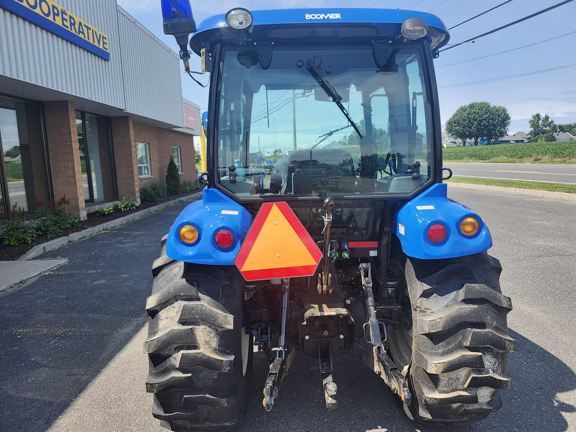 Un tracteur bleu est garé devant un bâtiment aux grandes fenêtres, affichant un panneau de sécurité triangulaire à l'arrière. Le ciel dégagé et la route sont visibles en arrière-plan.