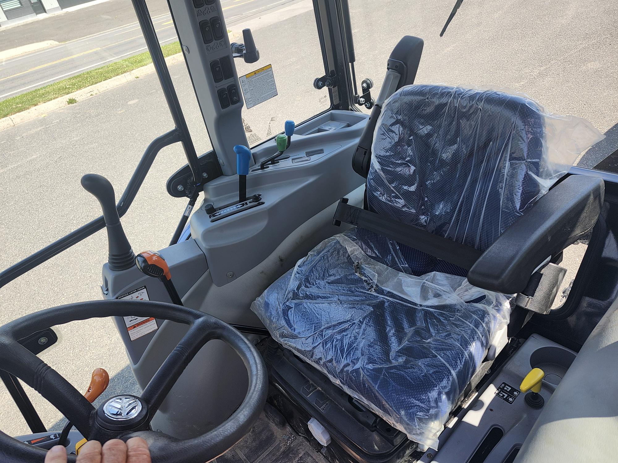 Vue de l'intérieur d'un tracteur, mettant en valeur le volant et un siège recouvert d'un plastique protecteur. Différents leviers et boutons de commande sont visibles, avec une vue partielle sur la route extérieure à travers le pare-brise.