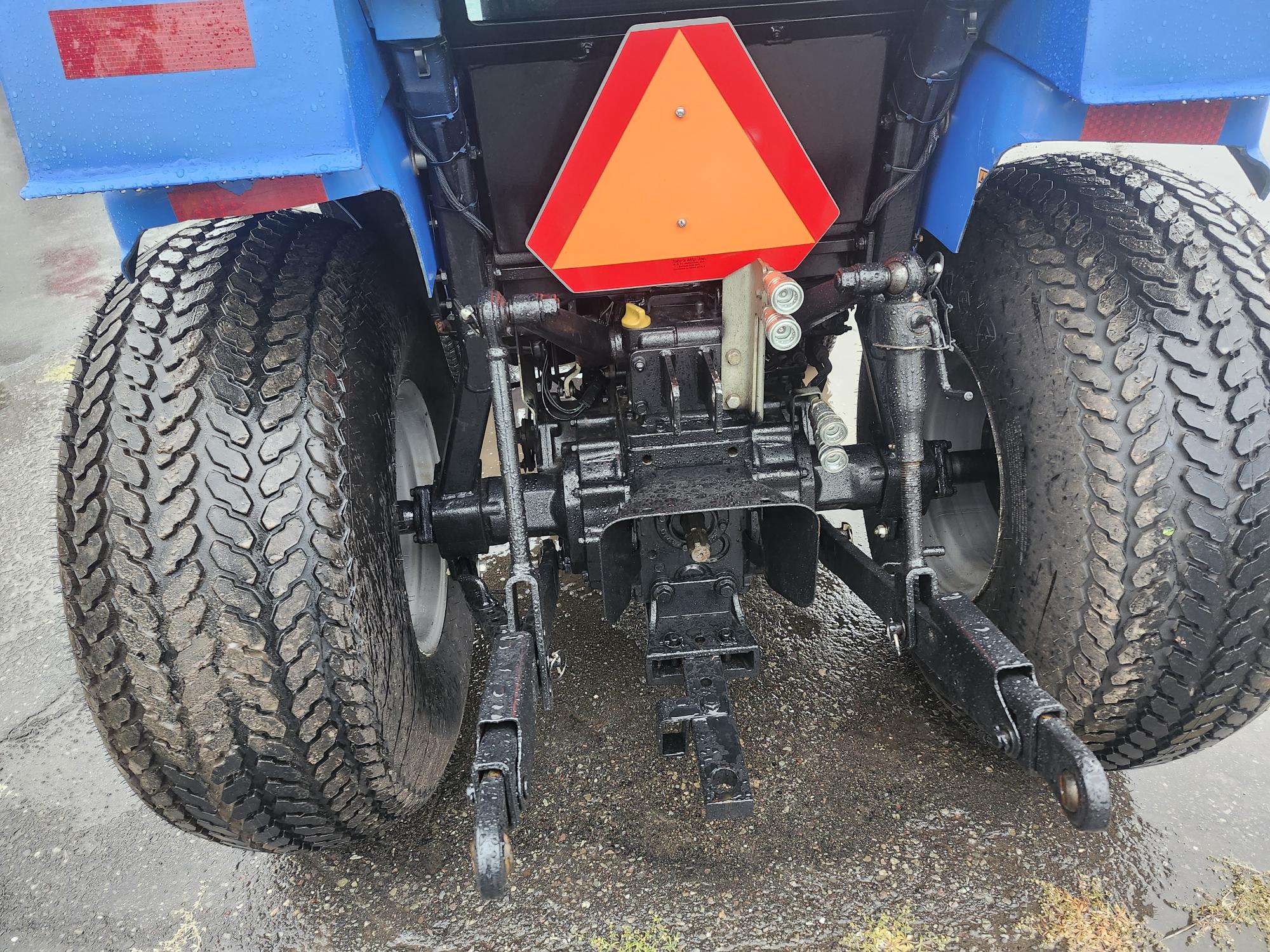 Gros plan de l'arrière d'un tracteur bleu, montrant les gros pneus texturés, un réflecteur triangulaire orange vif et divers composants mécaniques. Le sol est humide, ce qui indique une pluie récente.