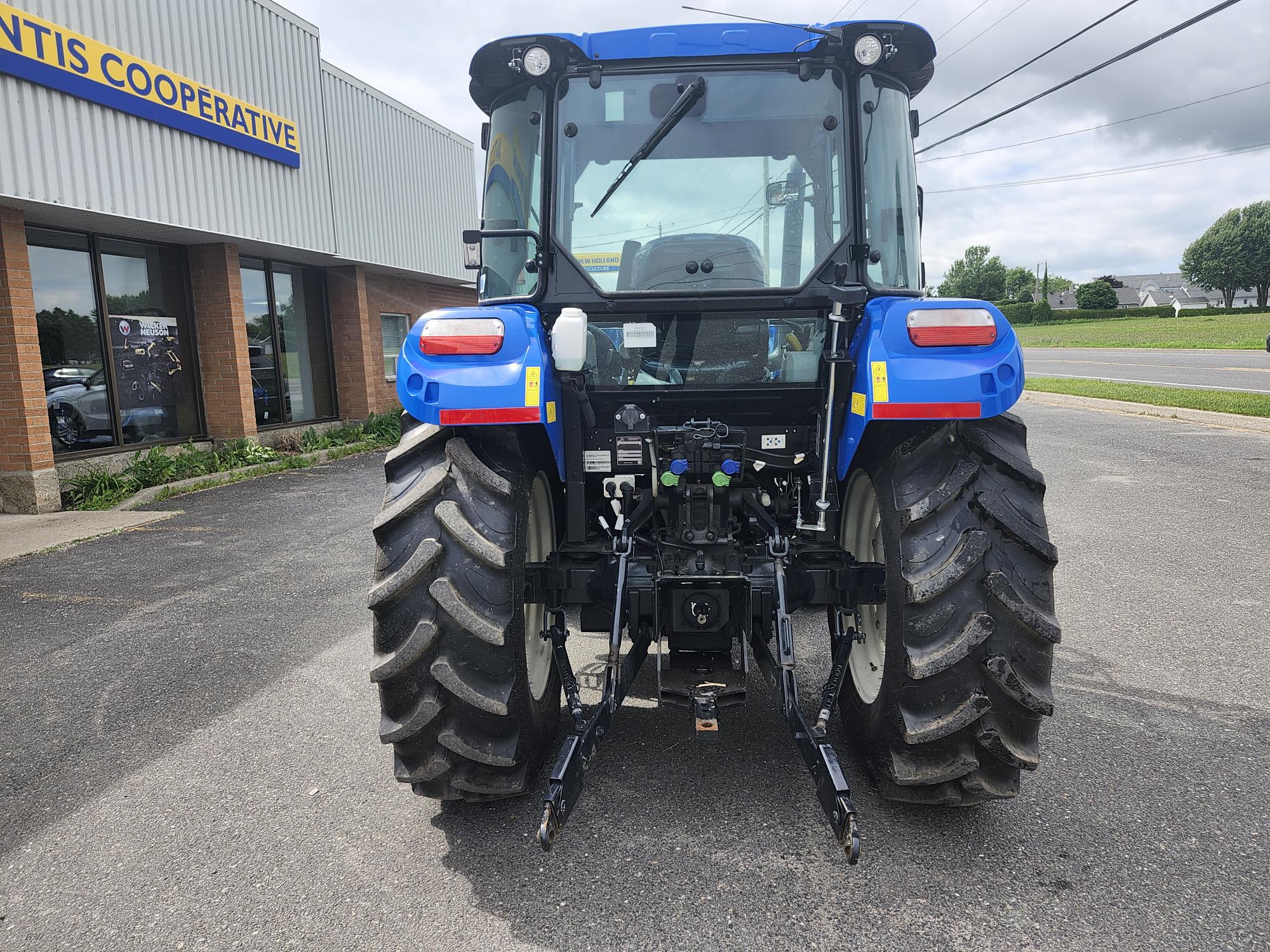 Vue arrière d'un tracteur bleu stationné sur une zone pavée devant un bâtiment. Le tracteur est équipé de gros pneus et de divers accessoires. Le bâtiment porte un panneau indiquant « COOPÉRATIVE » et il y a une zone herbeuse et une route à proximité.