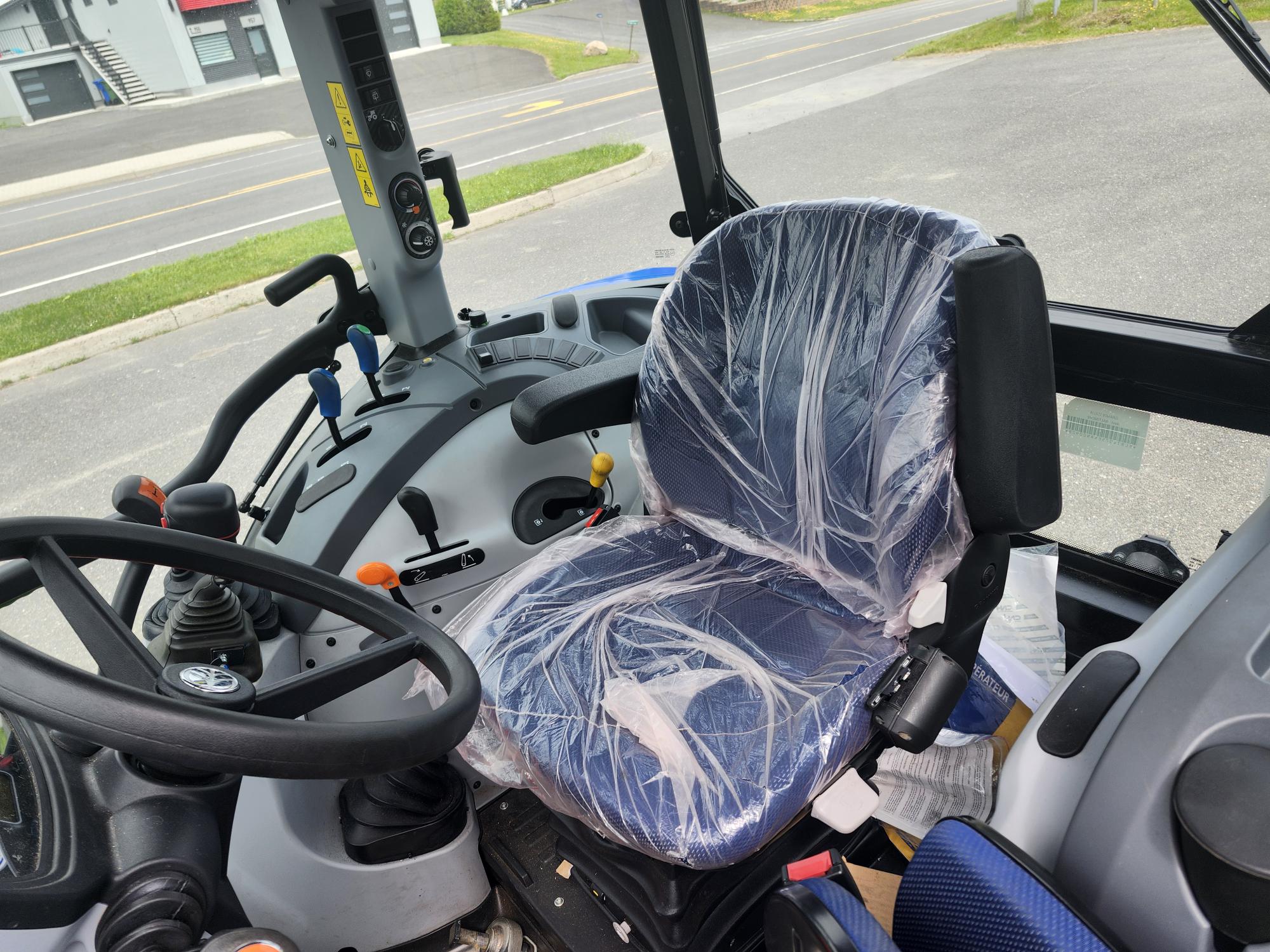 Intérieur d'une cabine de tracteur montrant le siège du conducteur enveloppé de plastique. Le volant est au premier plan, avec divers leviers de commande et boutons autour. Une route et de l'herbe sont visibles à l'extérieur des fenêtres.