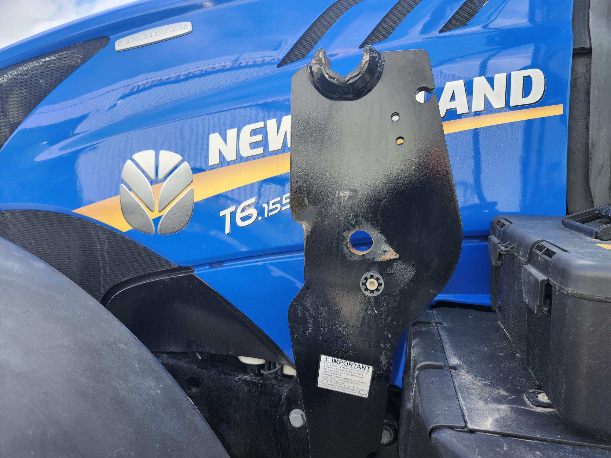 Gros plan d'un tracteur New Holland T6 155 bleu avec logo visible. Un composant métallique noir est au premier plan, avec une petite étiquette indiquant « Important ». Une boîte à outils est partiellement visible sur le côté droit.