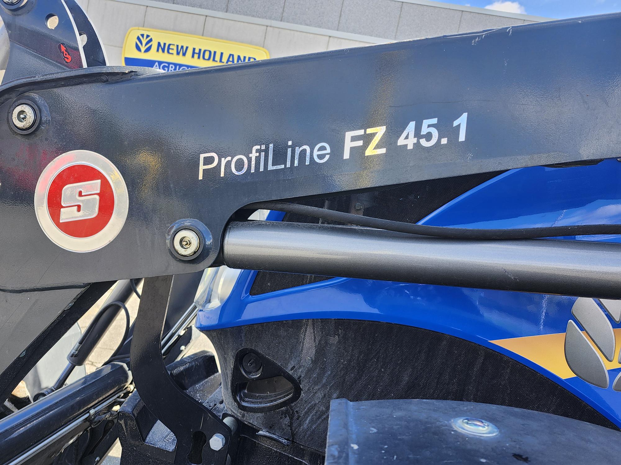 Gros plan d'une machine agricole sur laquelle est apposé le logo du modèle « ProfiLine FZ 45.1 » sur un bras en métal noir. L'arrière-plan comprend un panneau New Holland Agriculture partiellement visible sur un bâtiment.