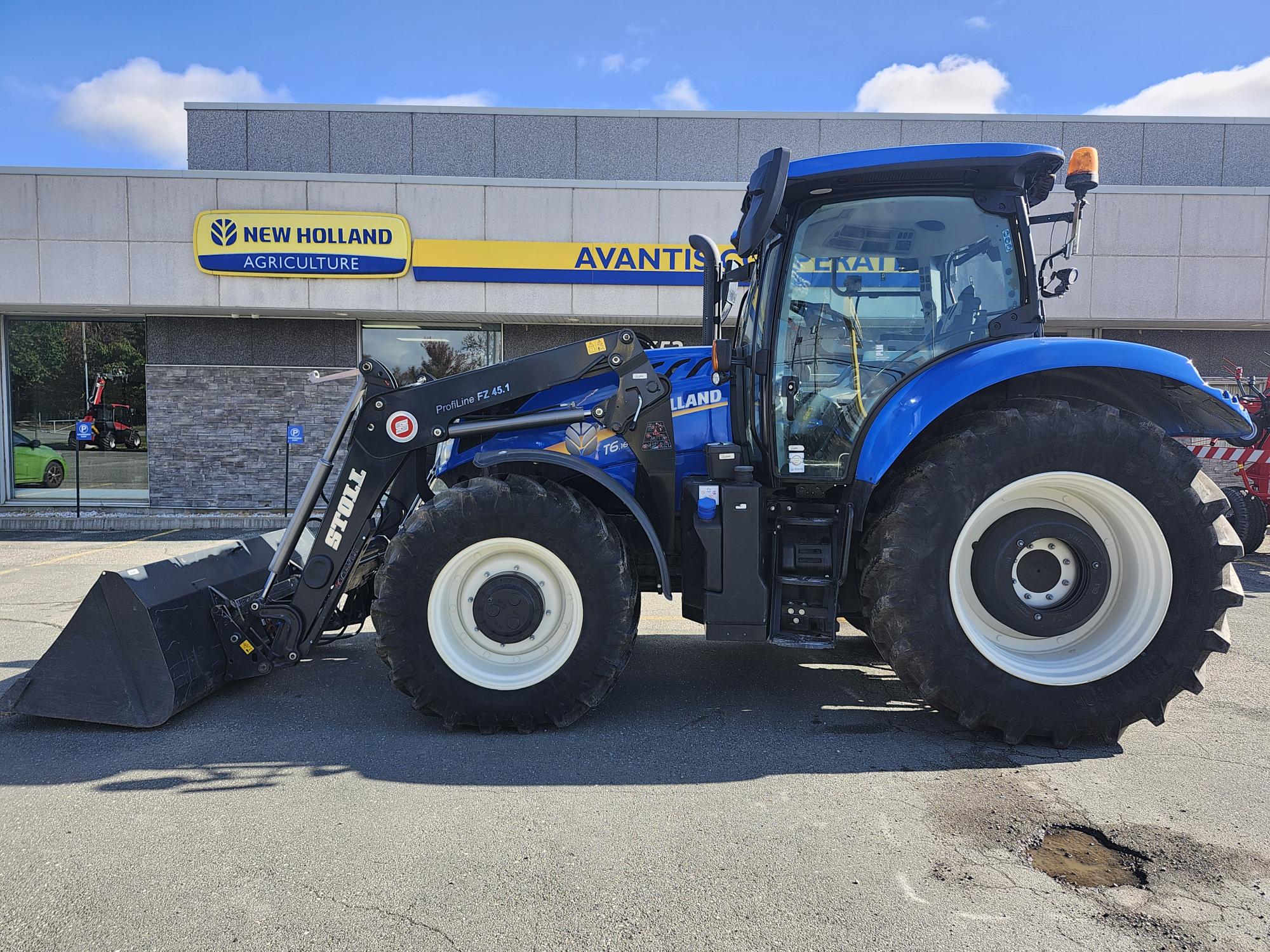 Un tracteur New Holland bleu équipé d'un chargeur frontal est garé devant un concessionnaire avec un panneau New Holland Agriculture. Le ciel est clair et ensoleillé.