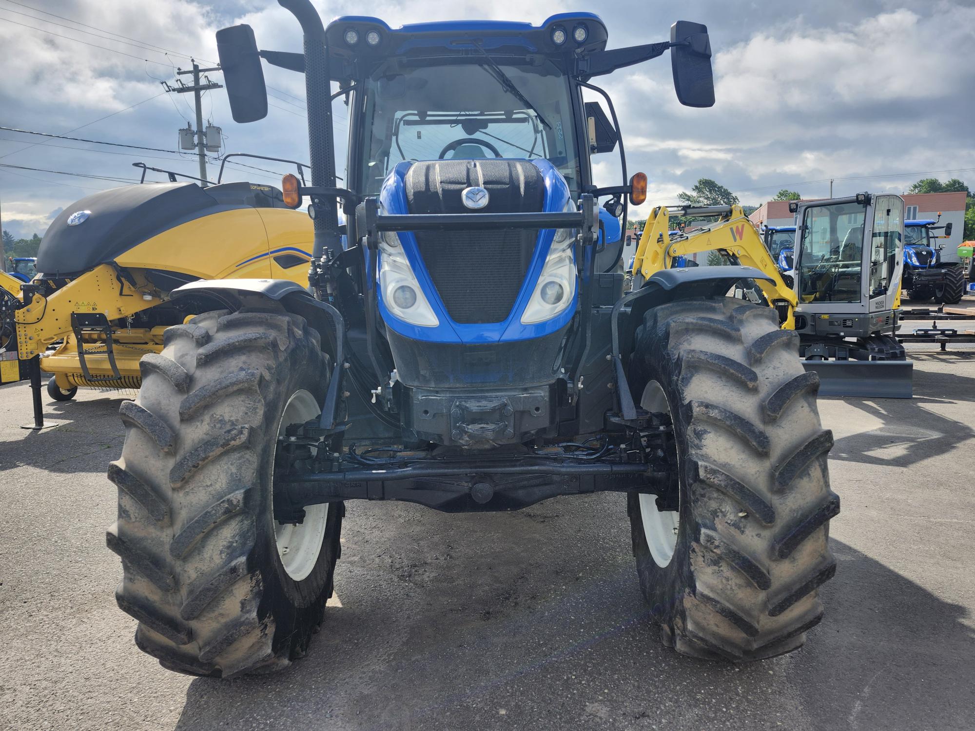 Un tracteur bleu et noir avec de gros pneus boueux est garé sur une surface en béton. Des engins de chantier, dont un véhicule jaune, sont visibles en arrière-plan sous un ciel nuageux.