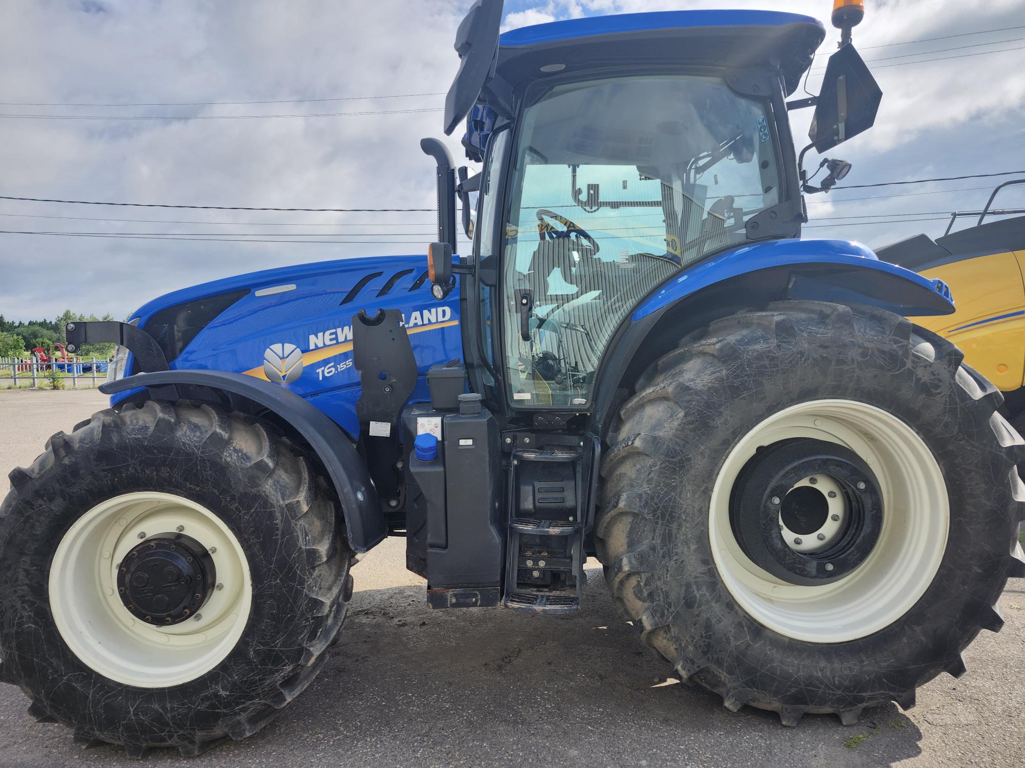 Un tracteur New Holland bleu est garé à l'extérieur par temps nuageux, mettant en valeur ses gros pneus et son design agricole.