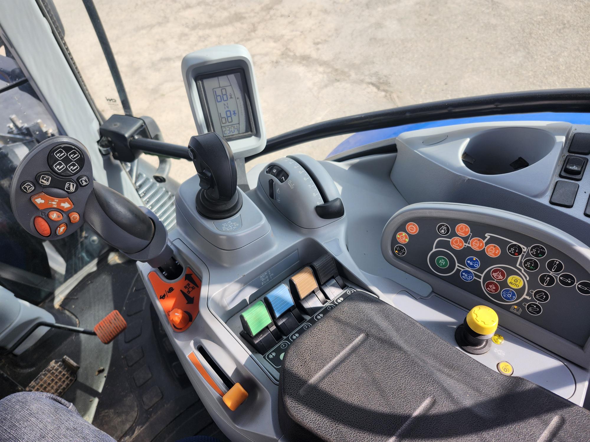 Vue intérieure d'une cabine de tracteur montrant le volant avec les boutons de commande, le levier de vitesse, divers interrupteurs et un écran d'affichage. Le tableau de bord est équipé de boutons colorés et d'un porte-gobelet, avec une pédale au sol.