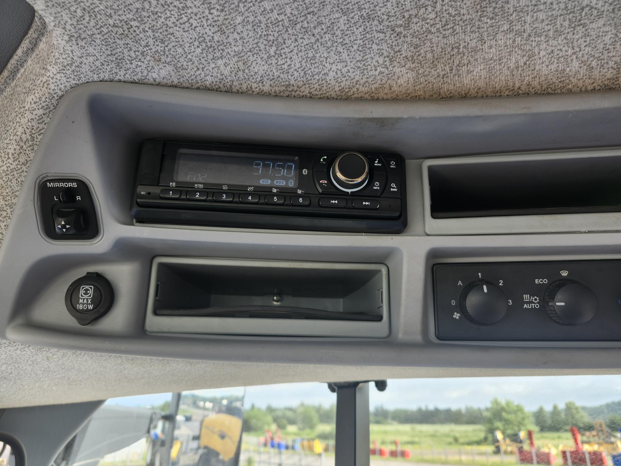 Vue intérieure du tableau de bord d'un véhicule montrant une radio réglée sur 97,5 FM, une commande de réglage des rétroviseurs et une bouche d'aération. Le tableau de bord est gris clair et un champ d'herbe avec des machines est visible à travers le pare-brise.