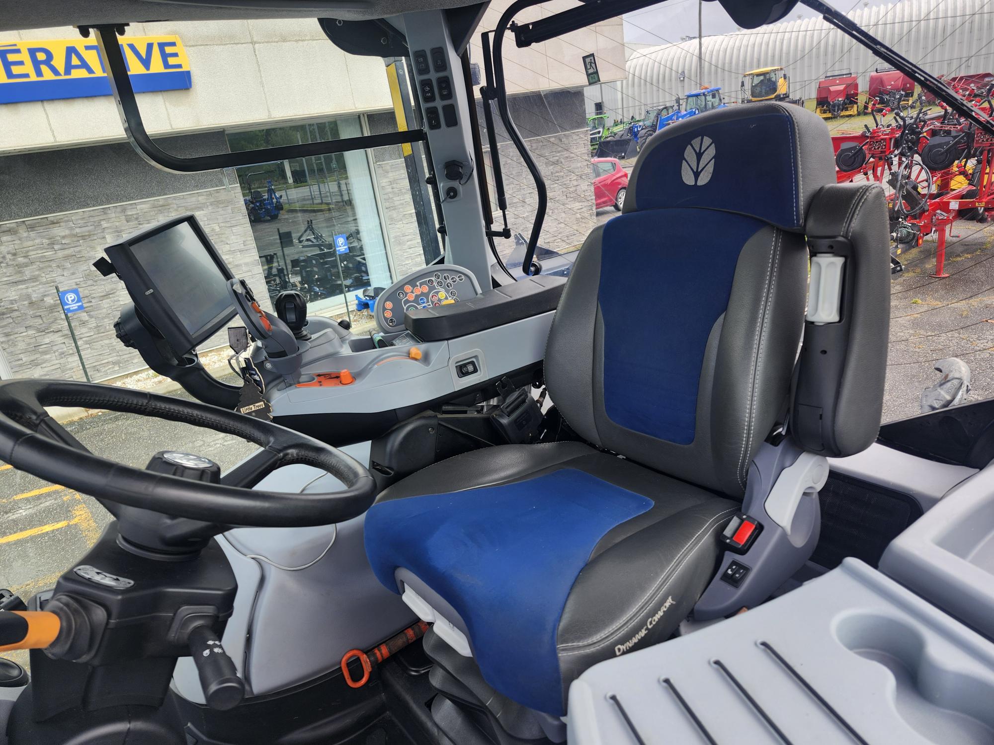 Vue intérieure de la cabine d'un tracteur moderne montrant un volant, un tableau de bord électronique avec écrans et un siège conducteur bleu et noir. L'arrière-plan présente une fenêtre donnant sur un équipement agricole stationné.