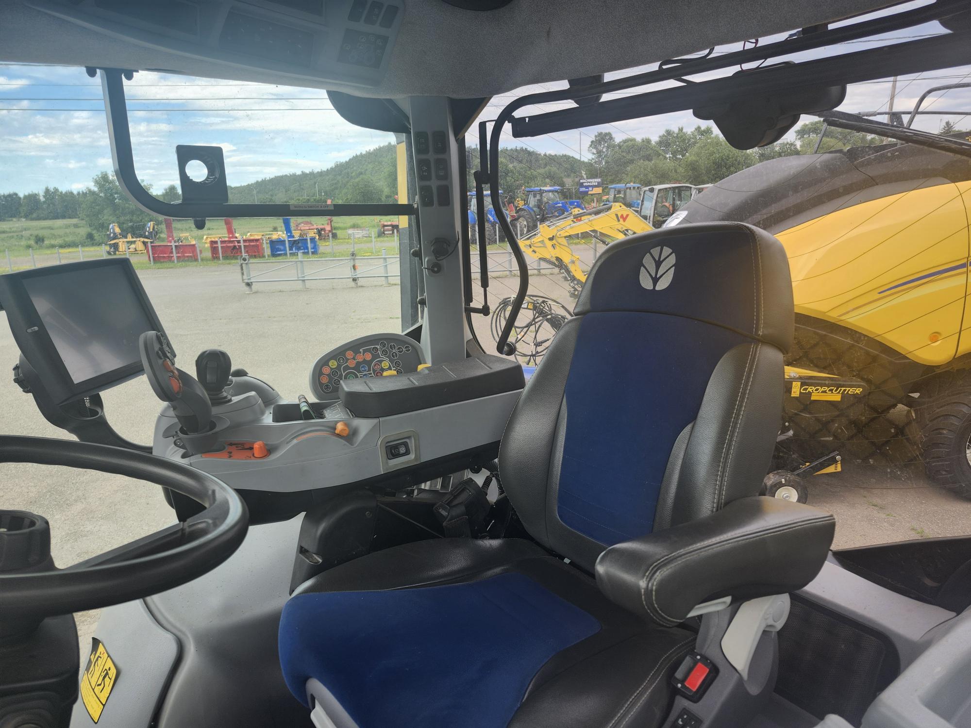 Vue intérieure d'une cabine de tracteur moderne montrant un volant, un panneau de commande avec divers boutons et un siège bleu. À travers la fenêtre, une vue floue sur l'équipement extérieur et la verdure est visible.