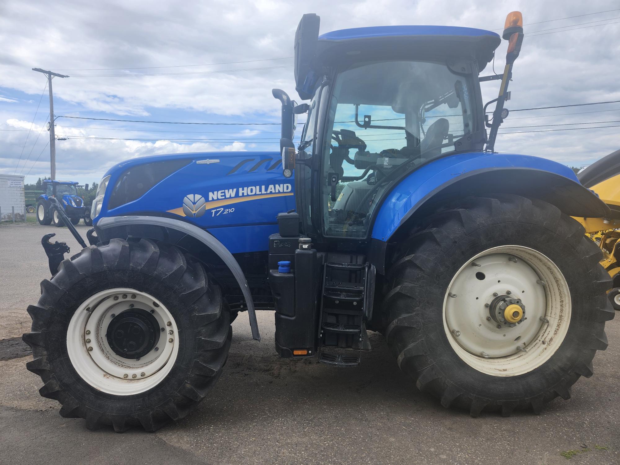 Un tracteur New Holland T7.290 bleu avec de gros pneus noirs est garé sur une surface pavée. Le ciel est nuageux en arrière-plan.