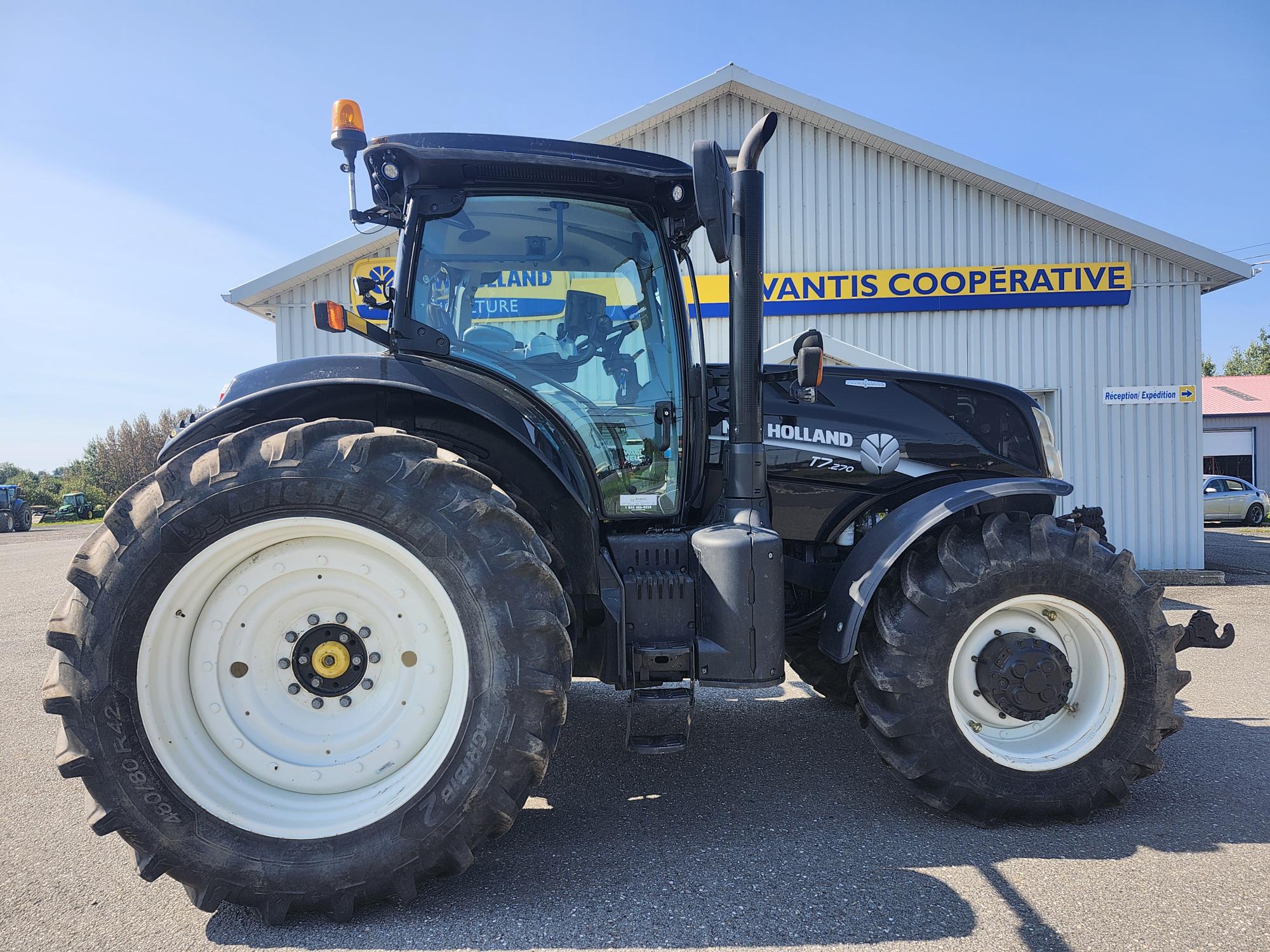 Un tracteur noir et bleu avec de grandes roues blanches est garé à l'extérieur d'un bâtiment portant l'inscription « Coopérative Advantis ». Le tracteur est doté d'une cabine compacte et est positionné sur une surface pavée sous un ciel bleu clair.