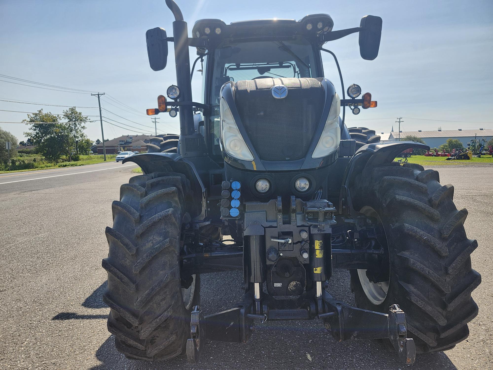 Un tracteur noir est garé sur une surface pavée sous un ciel bleu clair. La vue de face met en valeur les gros pneus et les fonctions utilitaires, avec des arbres et des poteaux électriques en arrière-plan.