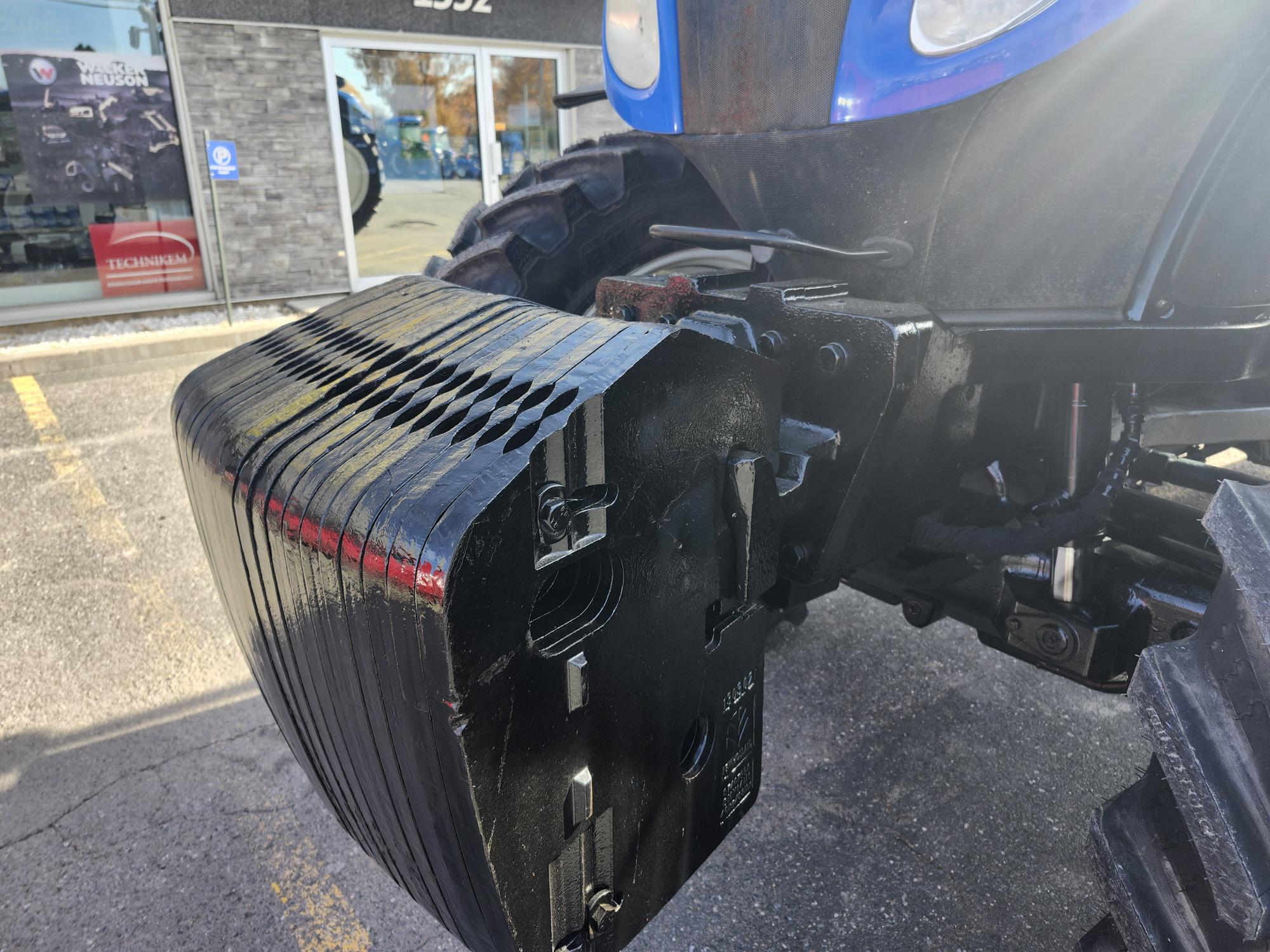 Gros plan d'un tracteur avec un focus sur les masses avant. Le tracteur est garé sur un parking, avec la lumière du soleil projetant des ombres et un magasin visible en arrière-plan. Un faible reflet du photographe est visible sur la surface du tracteur.