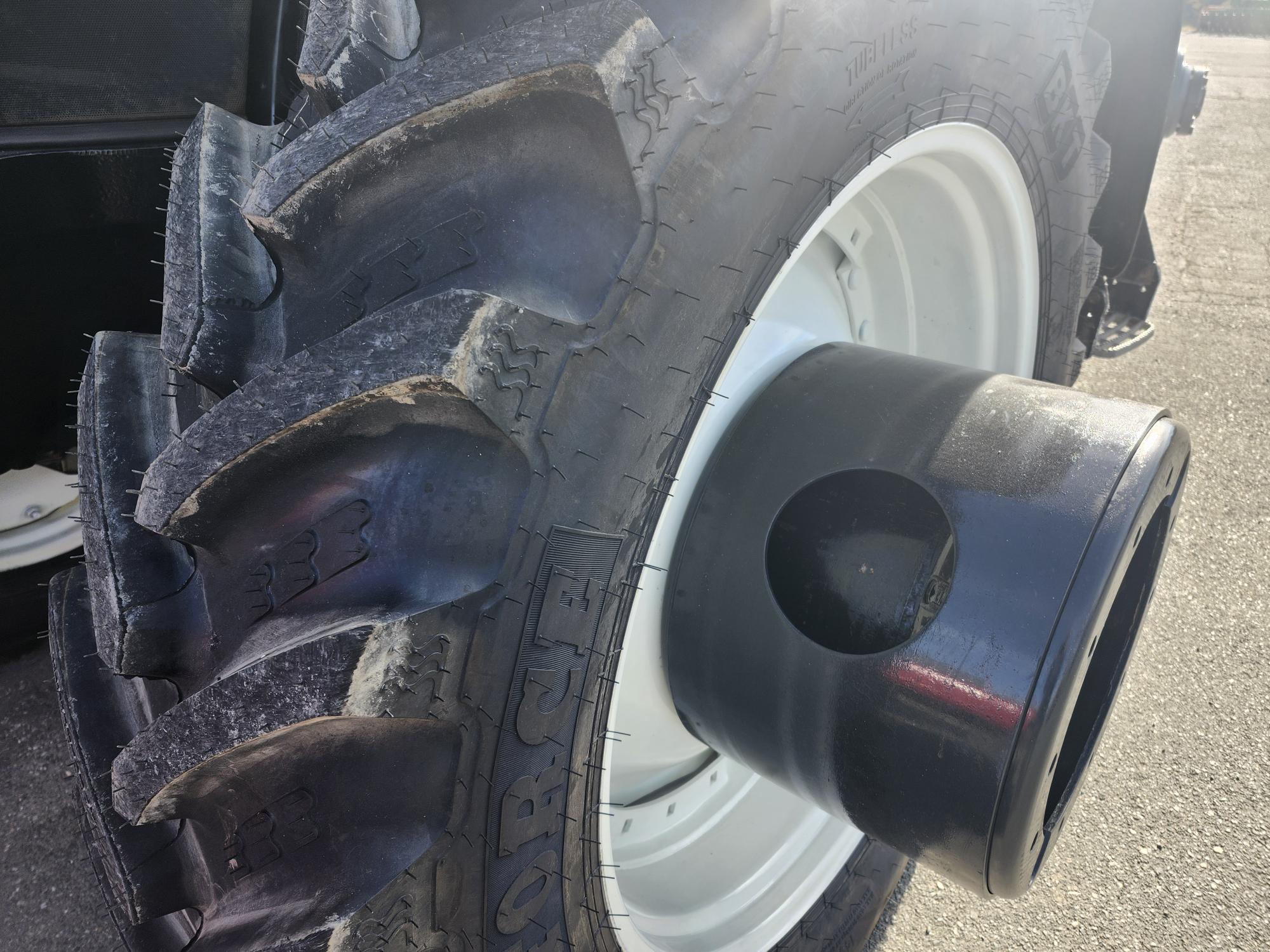 Gros plan d'un gros pneu de tracteur aux sculptures profondes sur un sol pavé. Le soleil brille, projetant des reflets sur la surface en caoutchouc. Un moyeu cylindrique noir est visible au centre de la roue.