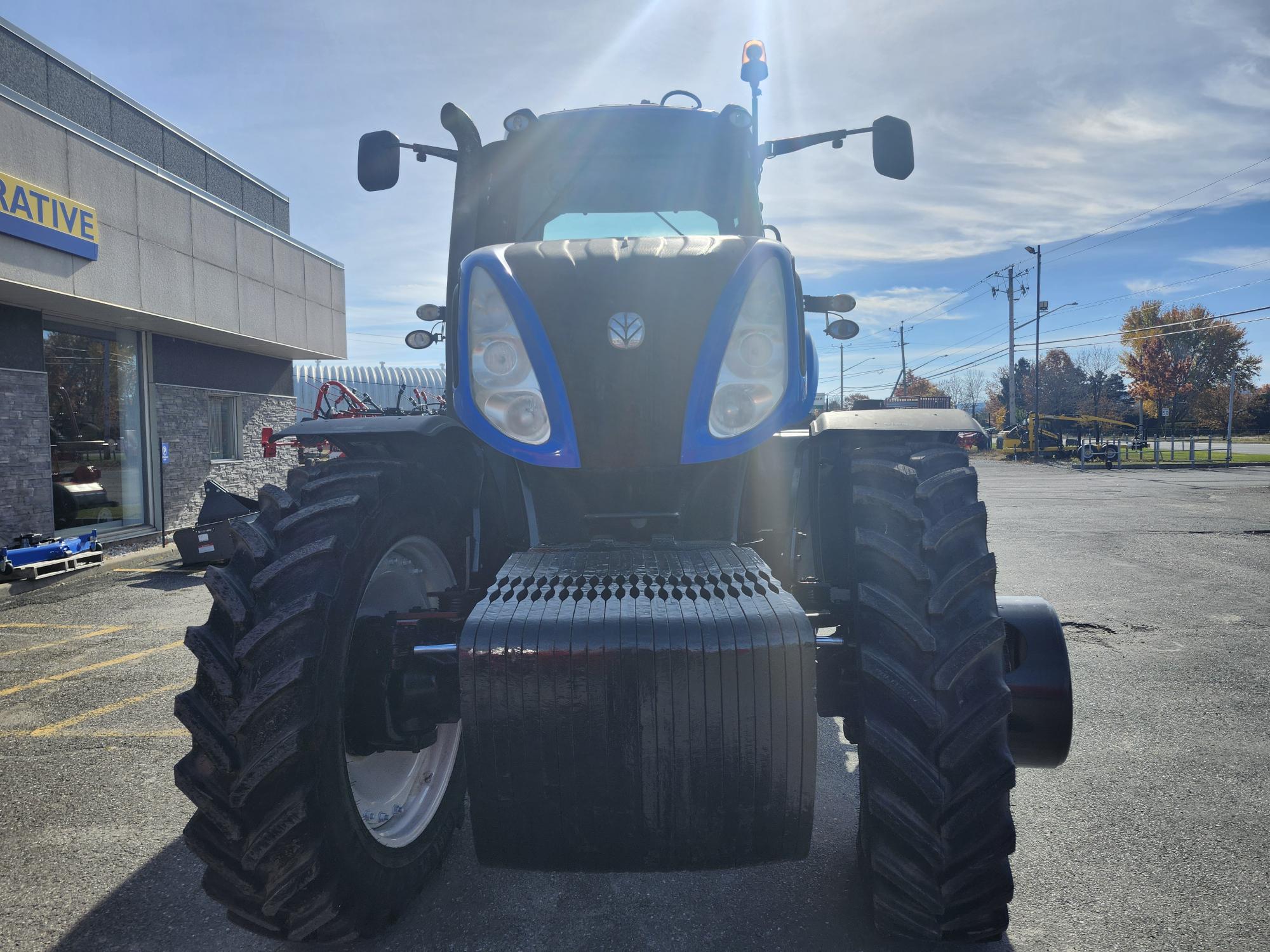 Vue de face d'un gros tracteur au design bleu et noir stationné sur un terrain pavé. Le soleil brille au-dessus, projetant des ombres. Le bâtiment et les lignes électriques sont visibles en arrière-plan.