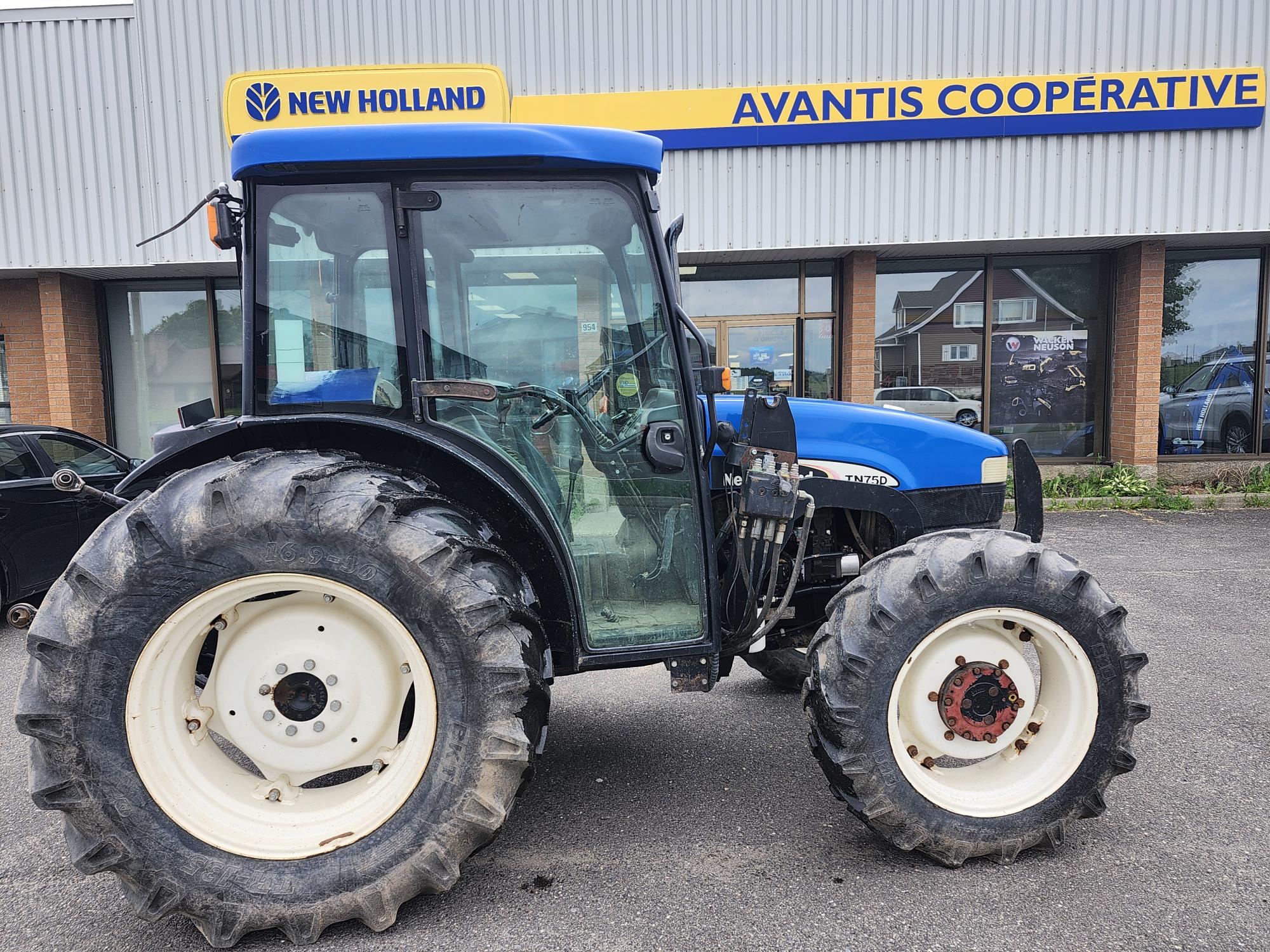 Un tracteur New Holland bleu est garé devant la coopérative Avantis. Le tracteur est présenté de profil, avec de gros pneus noirs et des jantes blanches. Le bâtiment en arrière-plan porte un panneau indiquant le nom de l'entreprise.