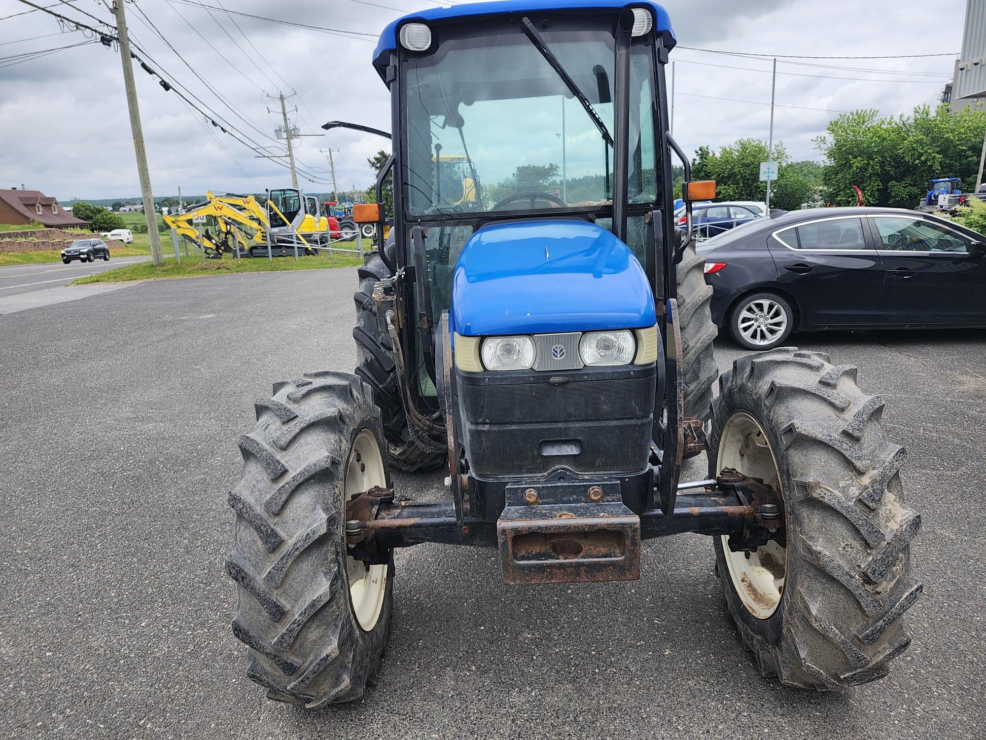 Un tracteur bleu avec de gros pneus est garé sur une surface pavée. En arrière-plan, on voit des voitures, des engins de chantier et des panneaux de signalisation sous un ciel nuageux.