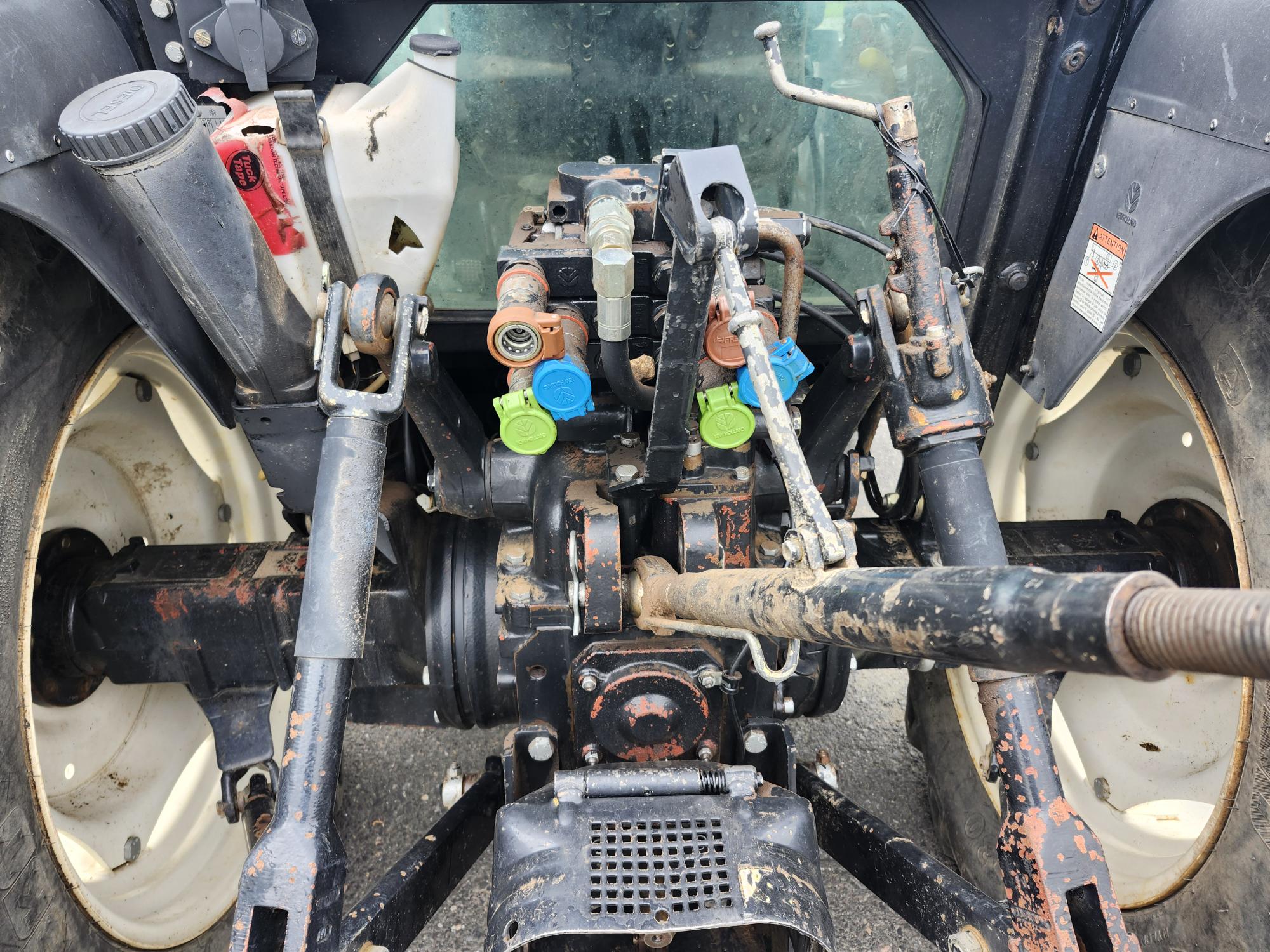 Gros plan de la partie arrière d'un tracteur, montrant des composants mécaniques, des connexions hydrauliques avec des bouchons à code couleur et des pièces robustes et patinées par les intempéries. Deux gros pneus arrière encadrent l'image.