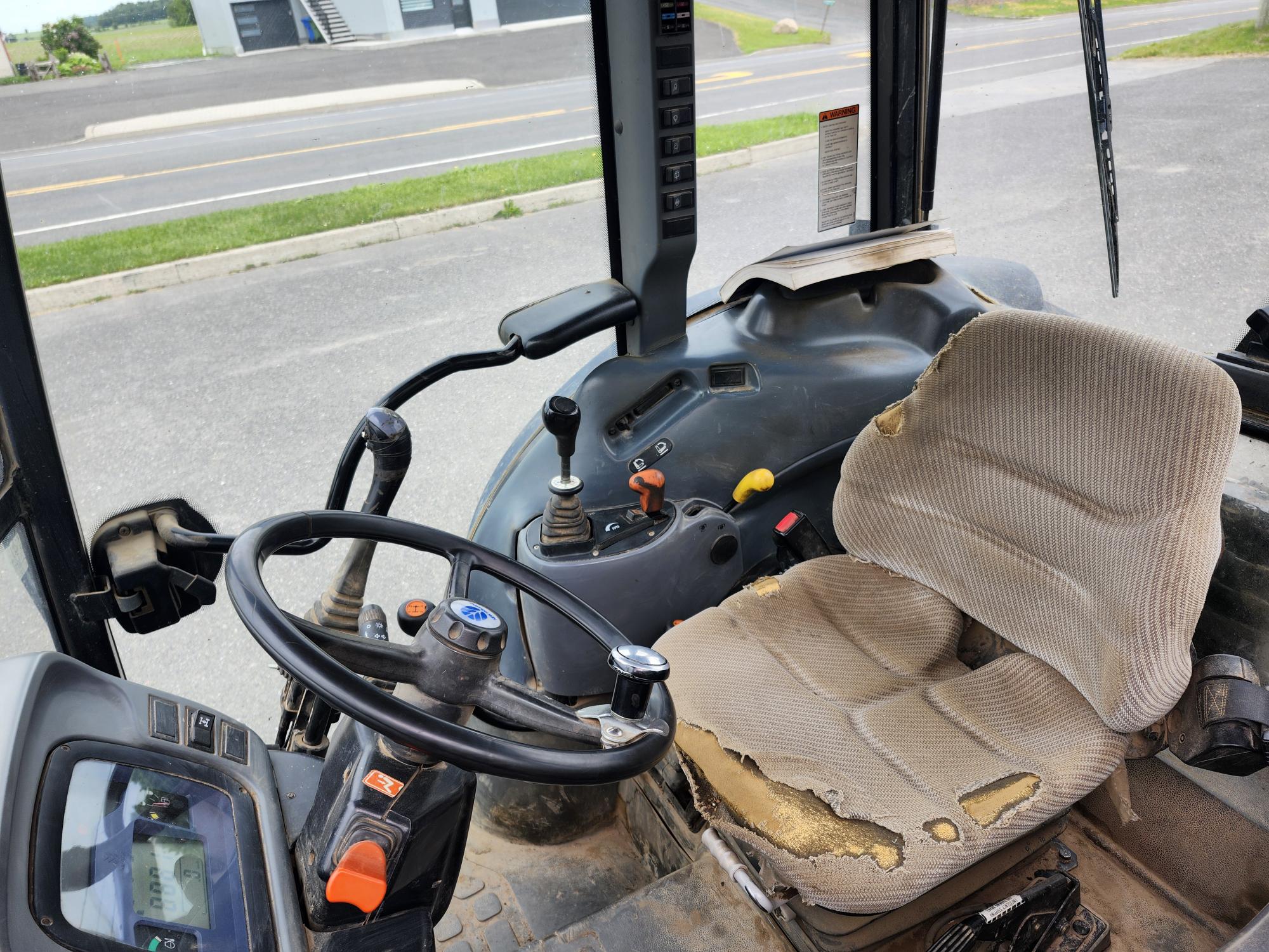 L'intérieur d'une cabine de tracteur usée avec volant, siège en tissu endommagé avec mousse apparente et leviers de commande. Un écran numérique est visible et une route est visible à l'extérieur à travers les fenêtres.