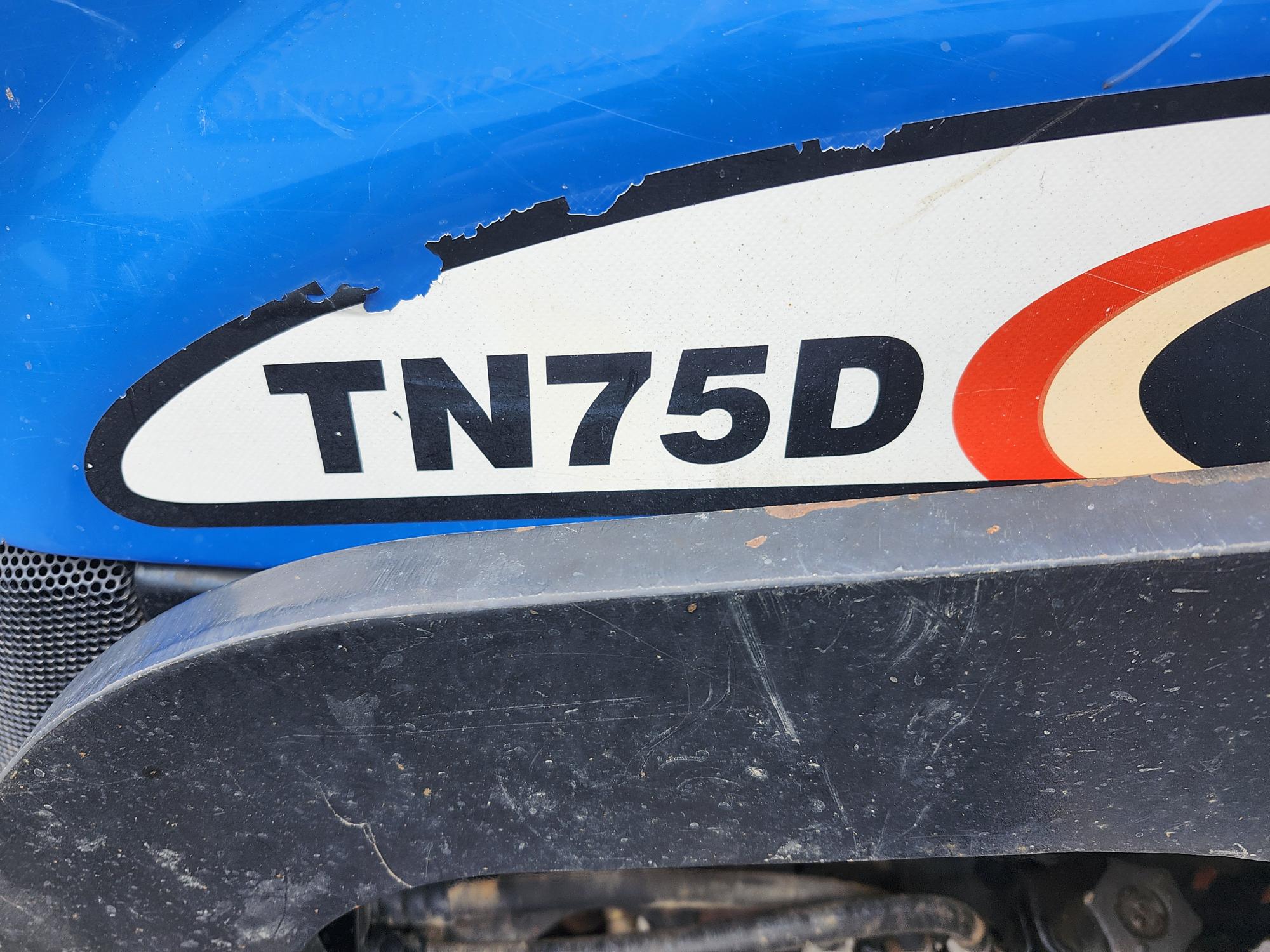 Gros plan d'un tracteur bleu sur lequel on peut voir un autocollant qui se décolle avec le texte « TN75D » en lettres noires en gras, à côté d'une ligne courbe rouge et orange. L'autocollant est placé au-dessus d'une surface métallique noire.