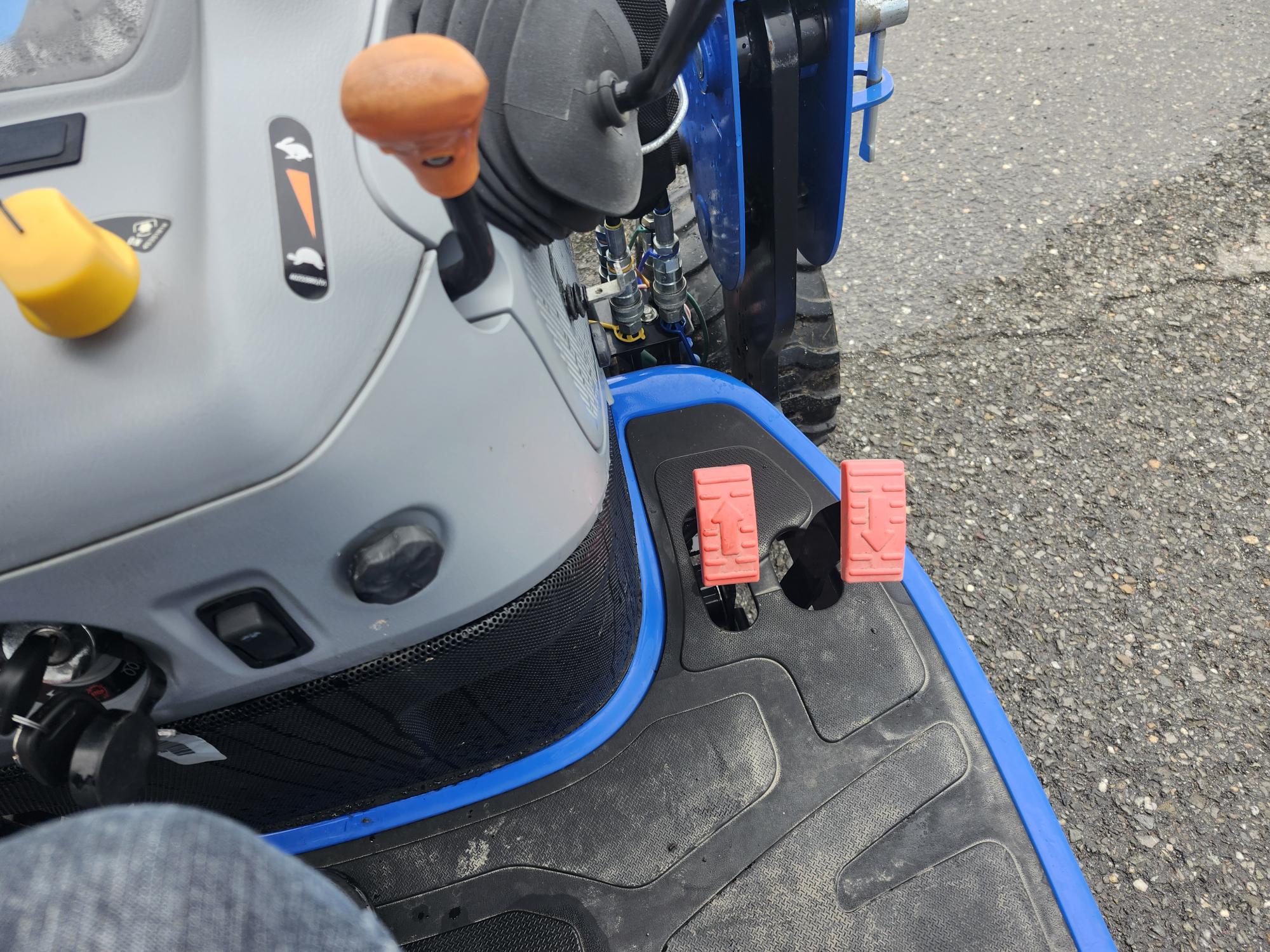 Gros plan des pédales d'un tracteur sur une surface texturée. Deux pédales rouges sont côte à côte, entourées d'un corps bleu et noir. Un volant et des leviers de commande sont partiellement visibles.