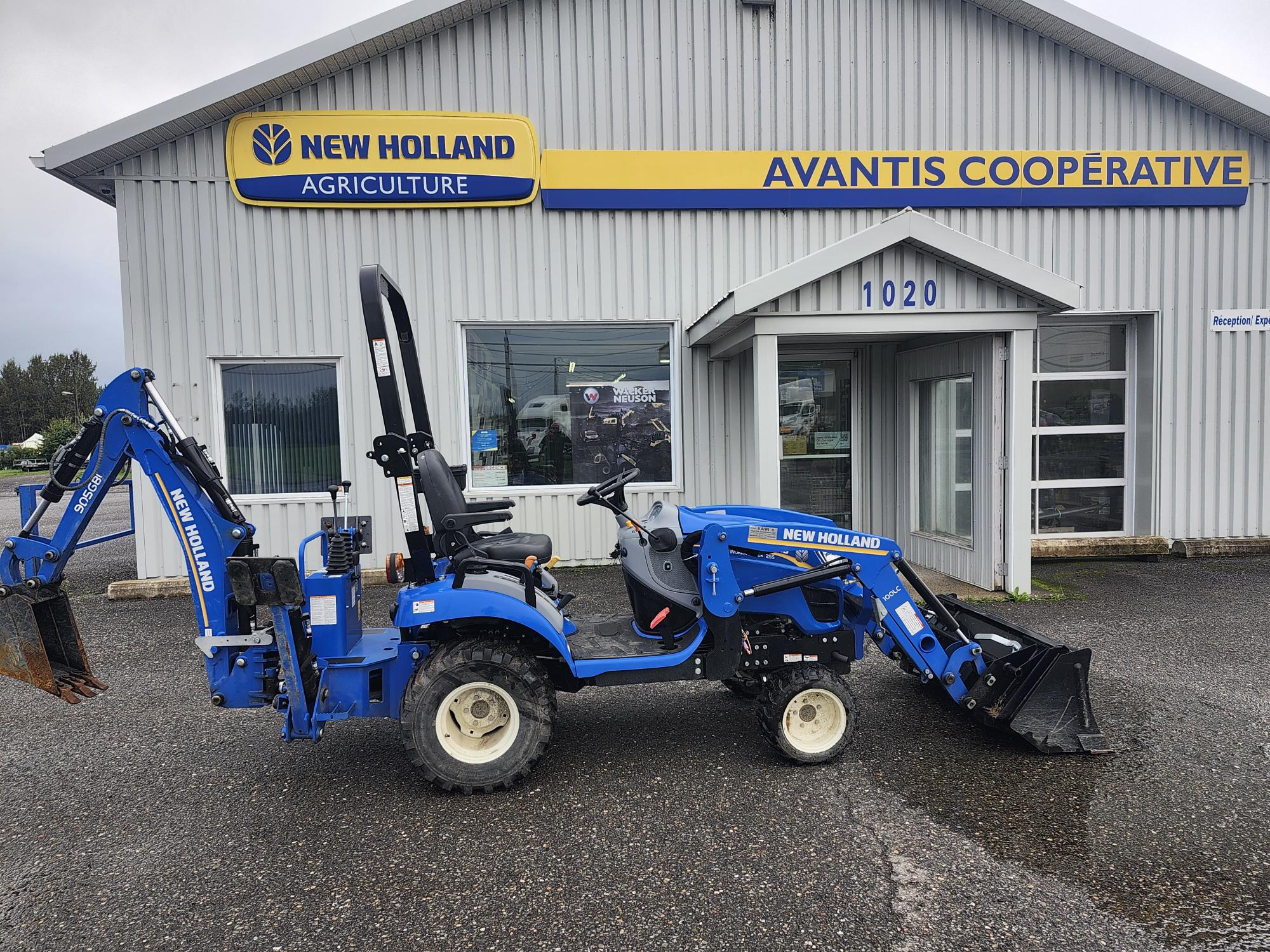 Un tracteur New Holland bleu équipé d'une rétrocaveuse et d'un chargeur frontal est garé devant Avantis Cooperative, un bâtiment de New Holland Agriculture. La scène est nuageuse, avec une surface pavée au premier plan.