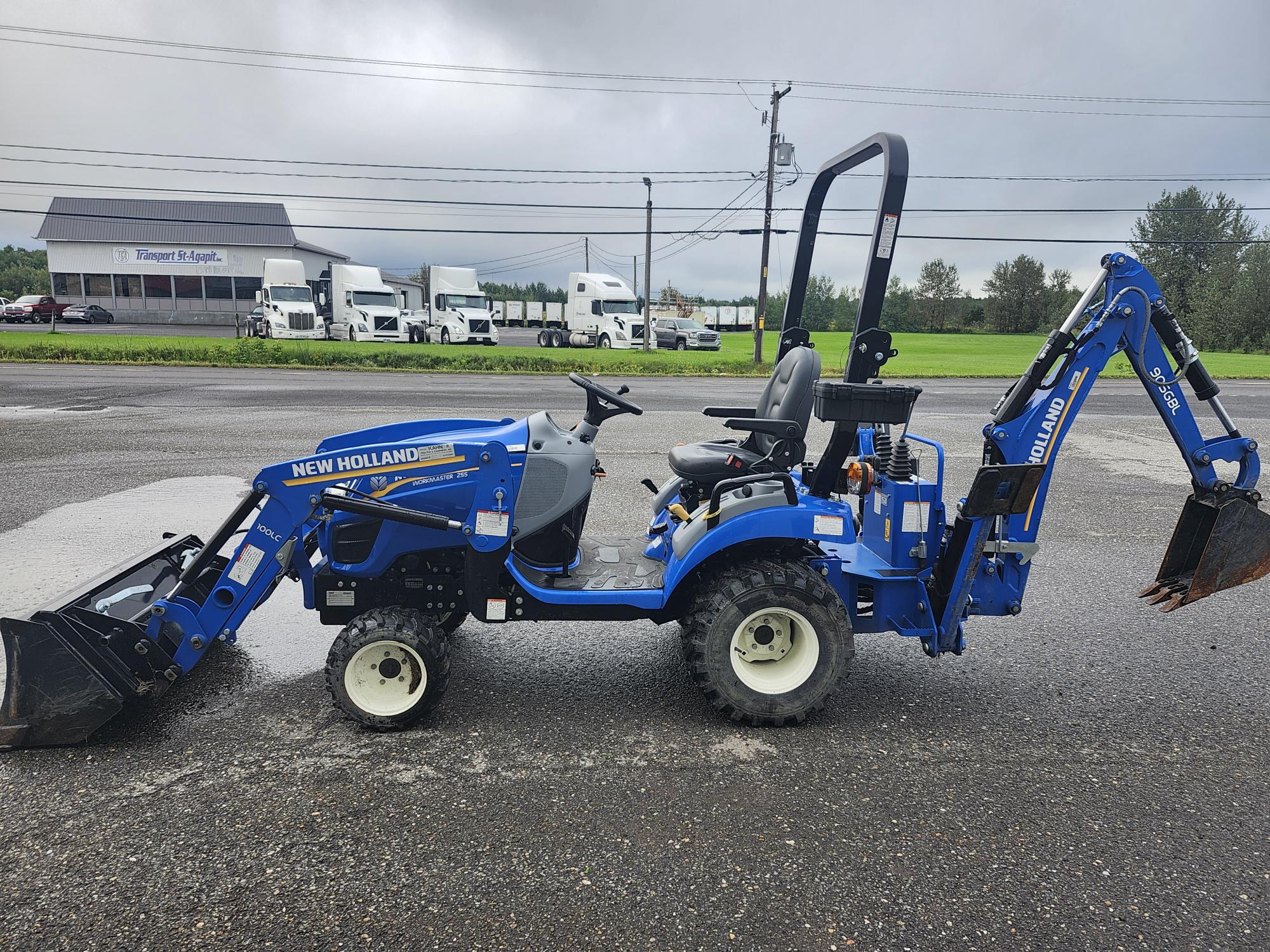 Un tracteur compact bleu New Holland équipé d'un chargeur frontal et d'une rétrocaveuse est stationné sur une surface asphaltée mouillée. À l'arrière-plan, on voit plusieurs camions blancs et un bâtiment avec le panneau « Finnegan's Superstore ».