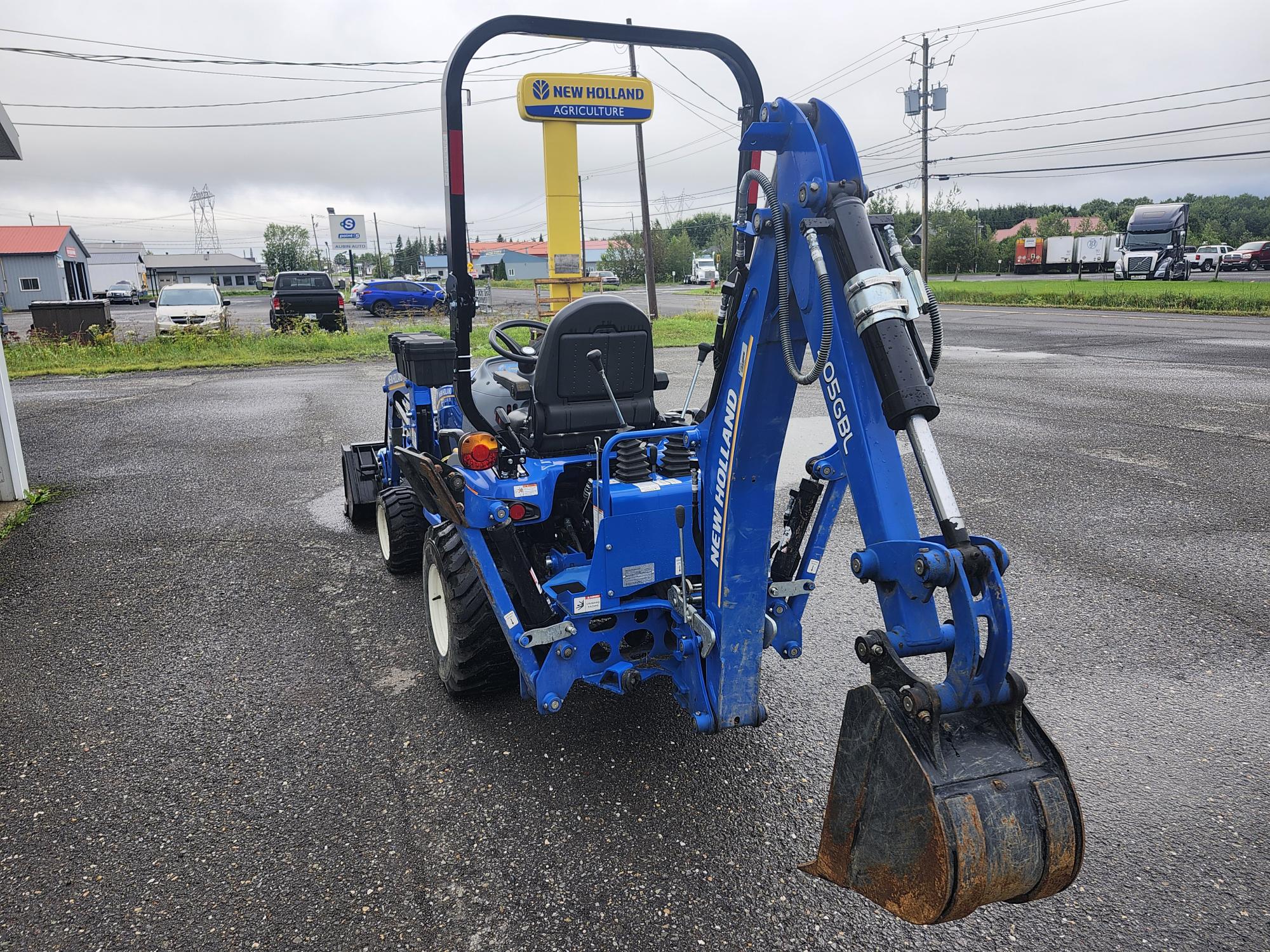 Une chargeuse-pelleteuse bleue est garée sur un terrain asphalté chez un concessionnaire New Holland. La machine est tournée dans le sens opposé à la caméra, avec des commandes visibles et un godet de creusement. Le ciel est couvert et divers véhicules sont visibles en arrière-plan.