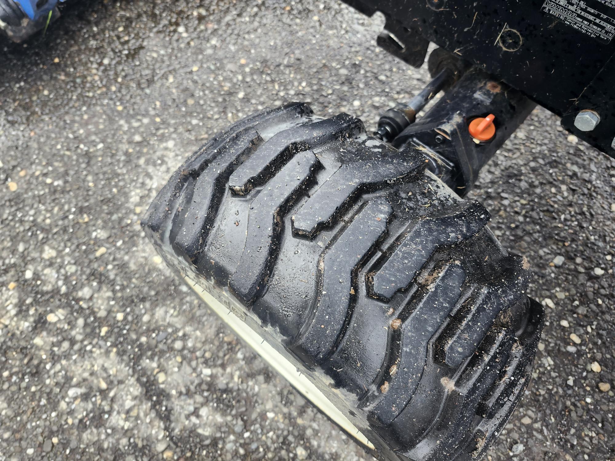 Gros plan d'un gros pneu noir robuste sur un véhicule, avec une bande de roulement détaillée. Le pneu est sur du gravier, ce qui suggère qu'il est adapté à une utilisation hors route ou intensive.