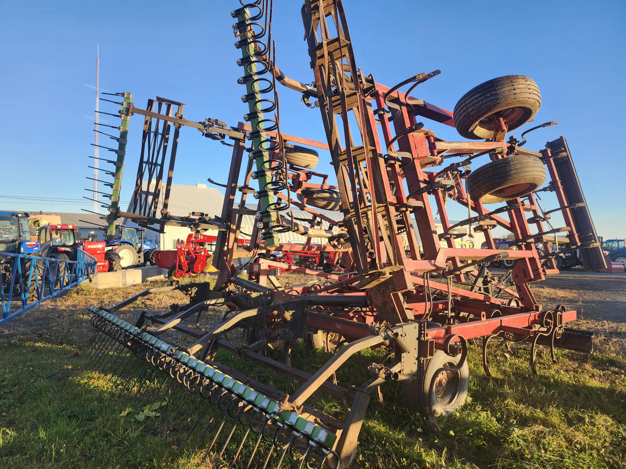 Un ensemble de machines agricoles de grande taille, dont des charrues et des herses, est garé sur un terrain herbeux sous un ciel bleu clair. Divers composants métalliques et roues sont visibles, avec un bâtiment et d'autres équipements en arrière-plan.