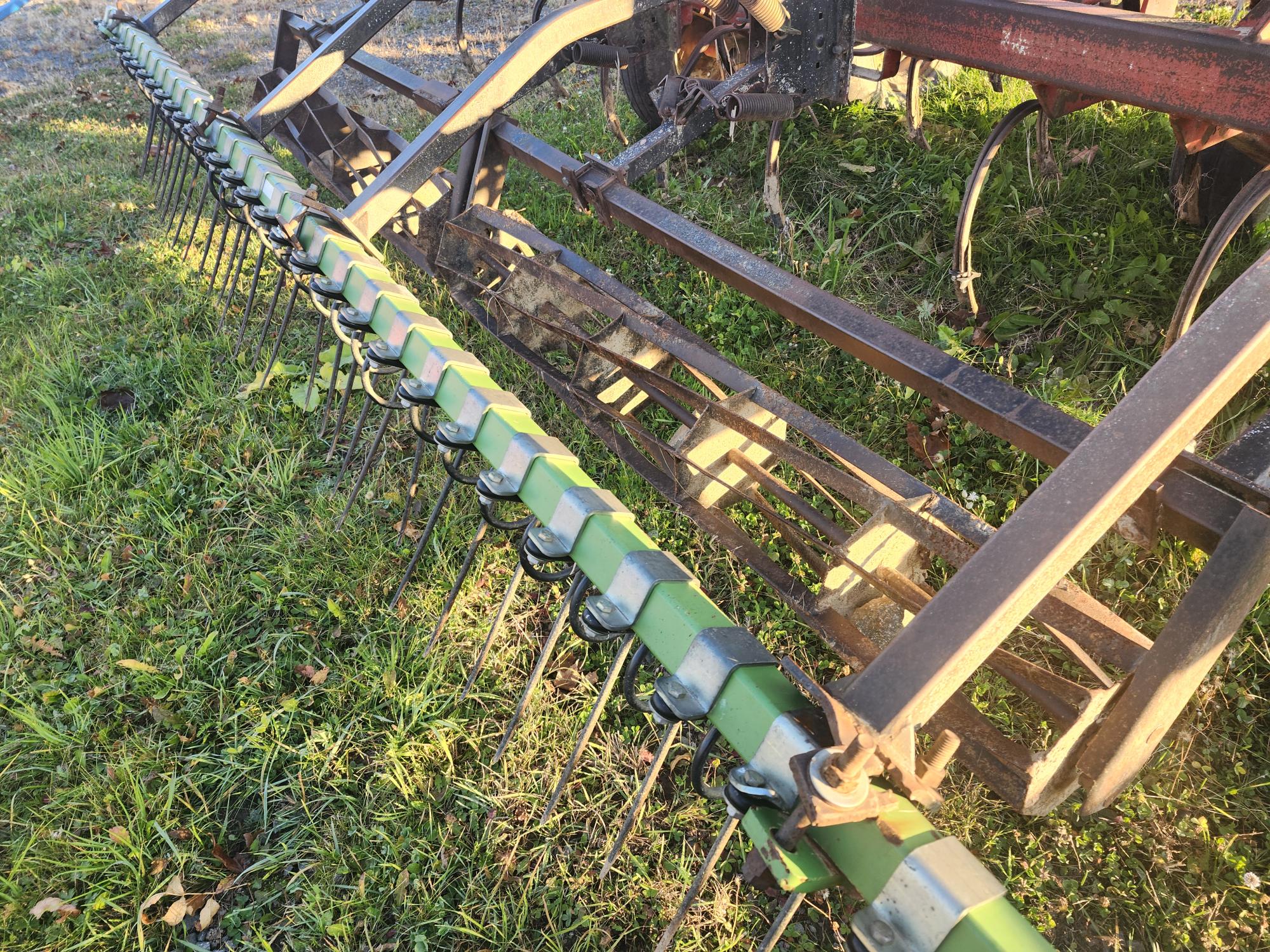 Gros plan d'une machine agricole rouillée dans un champ herbeux. Les composants métalliques comprennent une série de dents et d'éléments verts, probablement une partie d'un râteau à foin ou d'un équipement agricole similaire. Le sol est couvert d'herbe verte et de feuilles éparses.