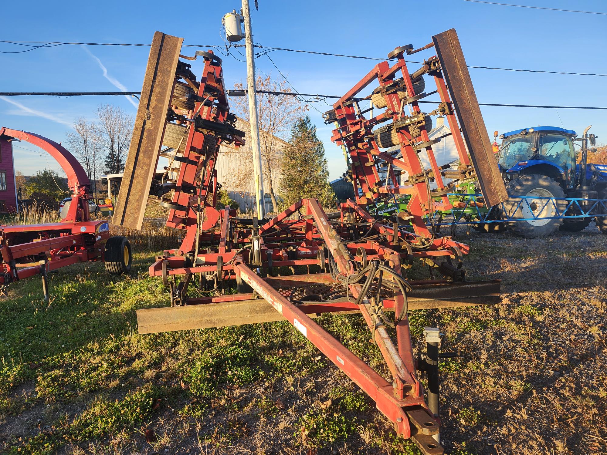 Une grande charrue agricole rouge à lames multiples et pièces en bois est garée sur un sol herbeux. Un tracteur bleu est visible en arrière-plan sous un ciel clair.