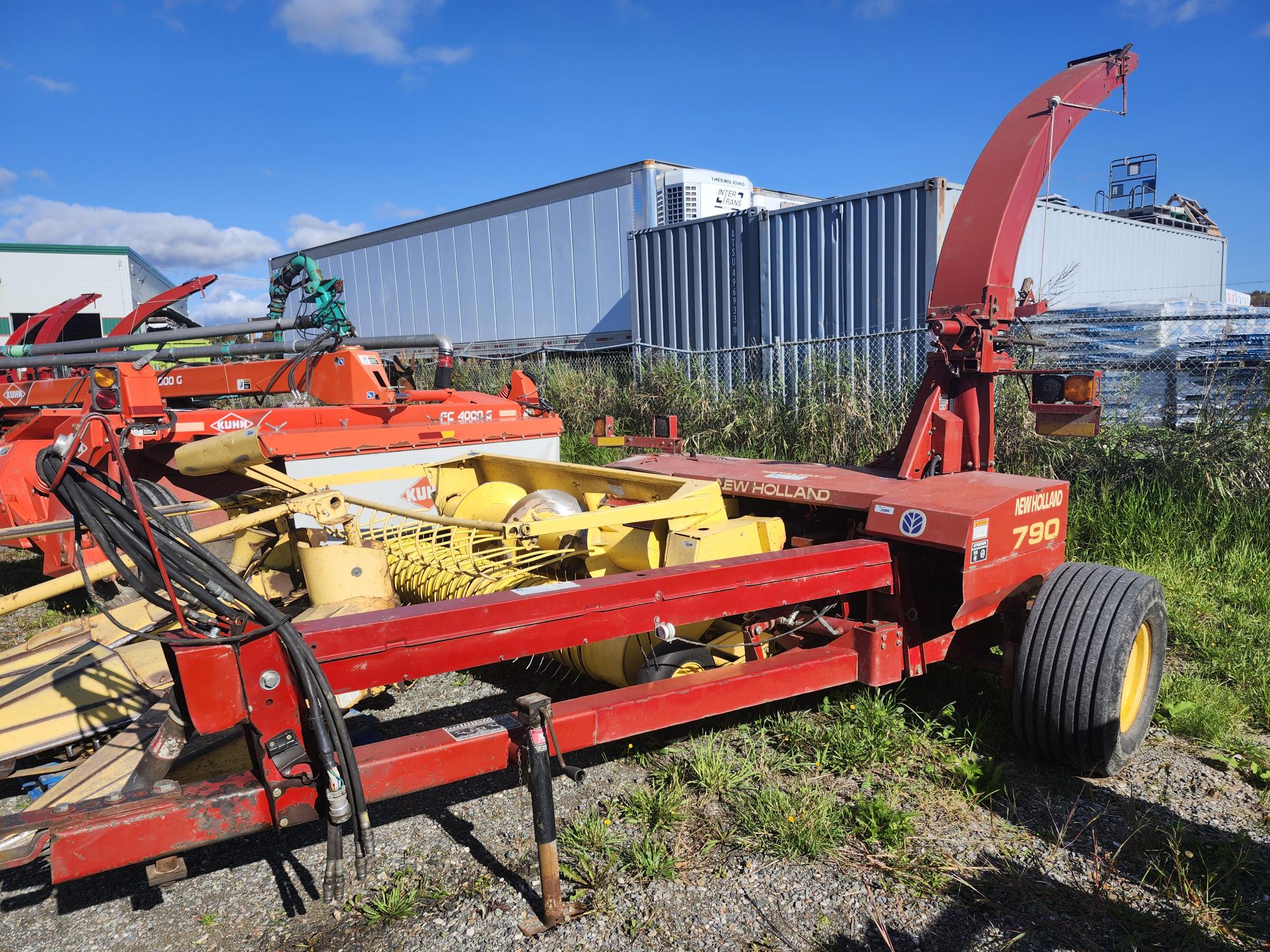 Une machine agricole rouge avec des composants jaunes, étiquetée « New Holland 790 », est garée sur l'herbe près d'une clôture métallique. L'arrière-plan présente des bâtiments industriels et un ciel bleu clair.