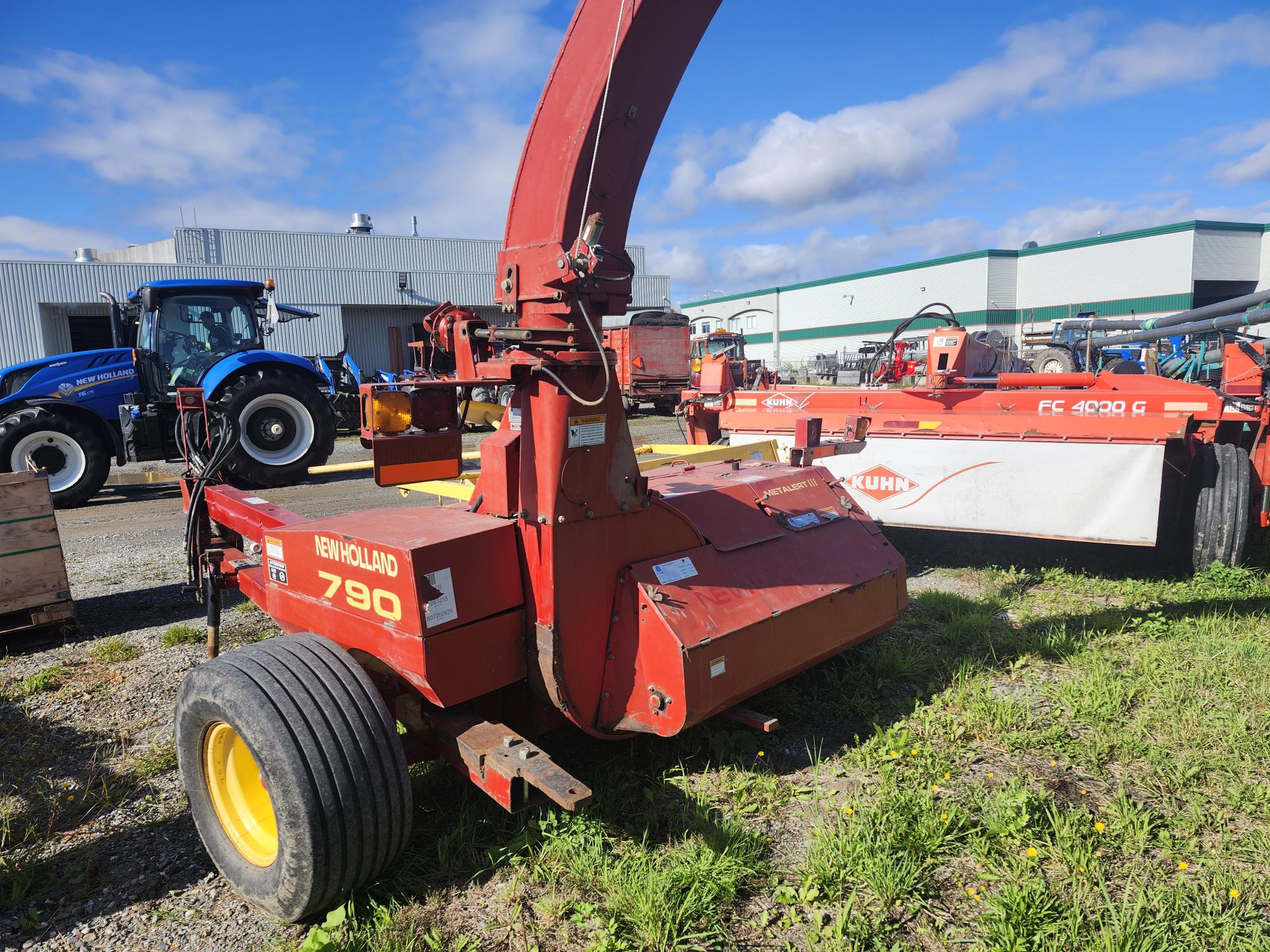 Une machine agricole New Holland 790 rouge à l'extérieur, avec un tracteur bleu et un autre outil agricole en arrière-plan. La scène comprend un entrepôt et un ciel partiellement nuageux.