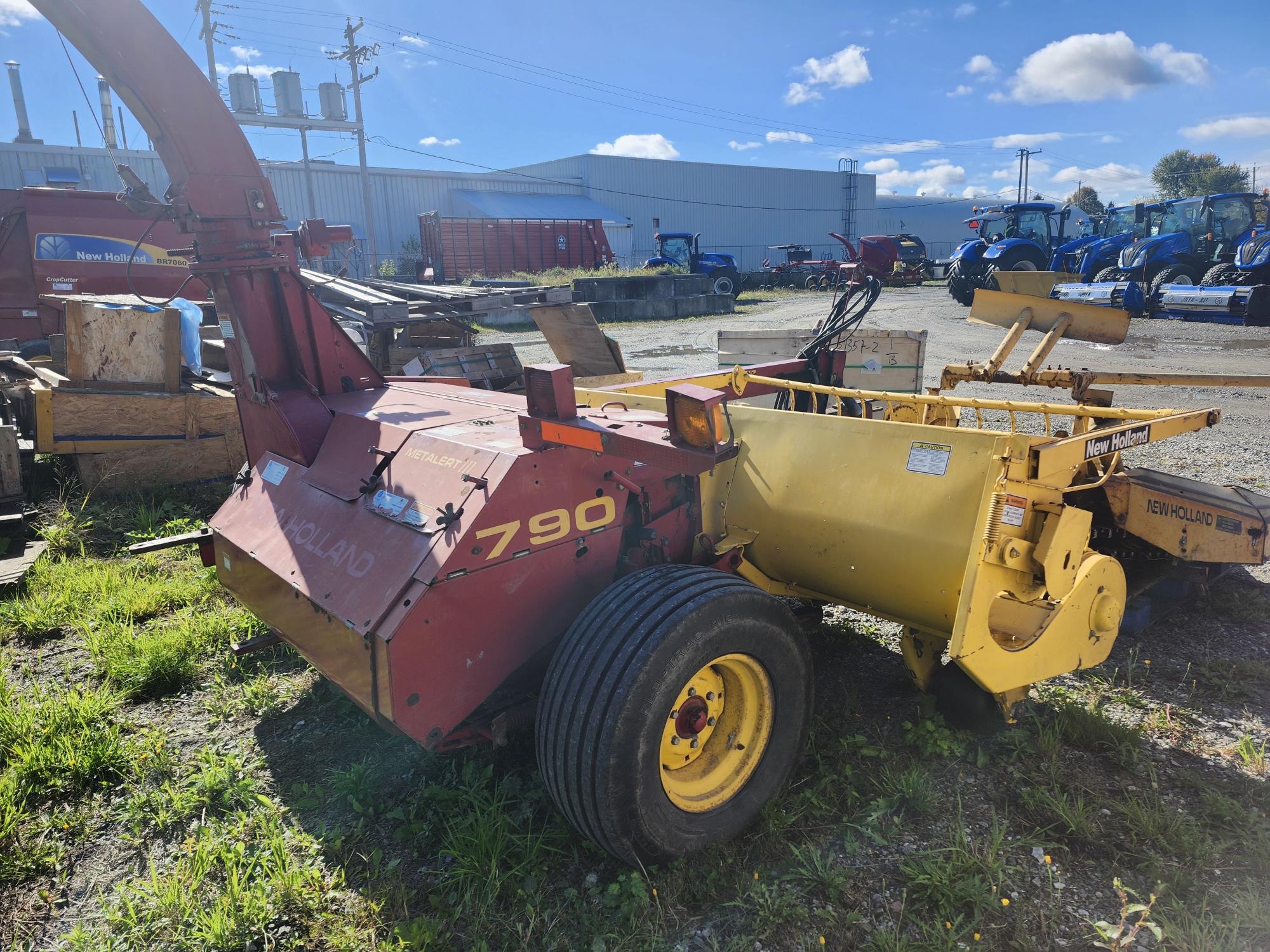 Une machine agricole New Holland 790 rouge et jaune est garée sur un terrain herbeux dans un parking extérieur. D'autres équipements agricoles et un bâtiment sont visibles à l'arrière-plan sous un ciel partiellement nuageux.