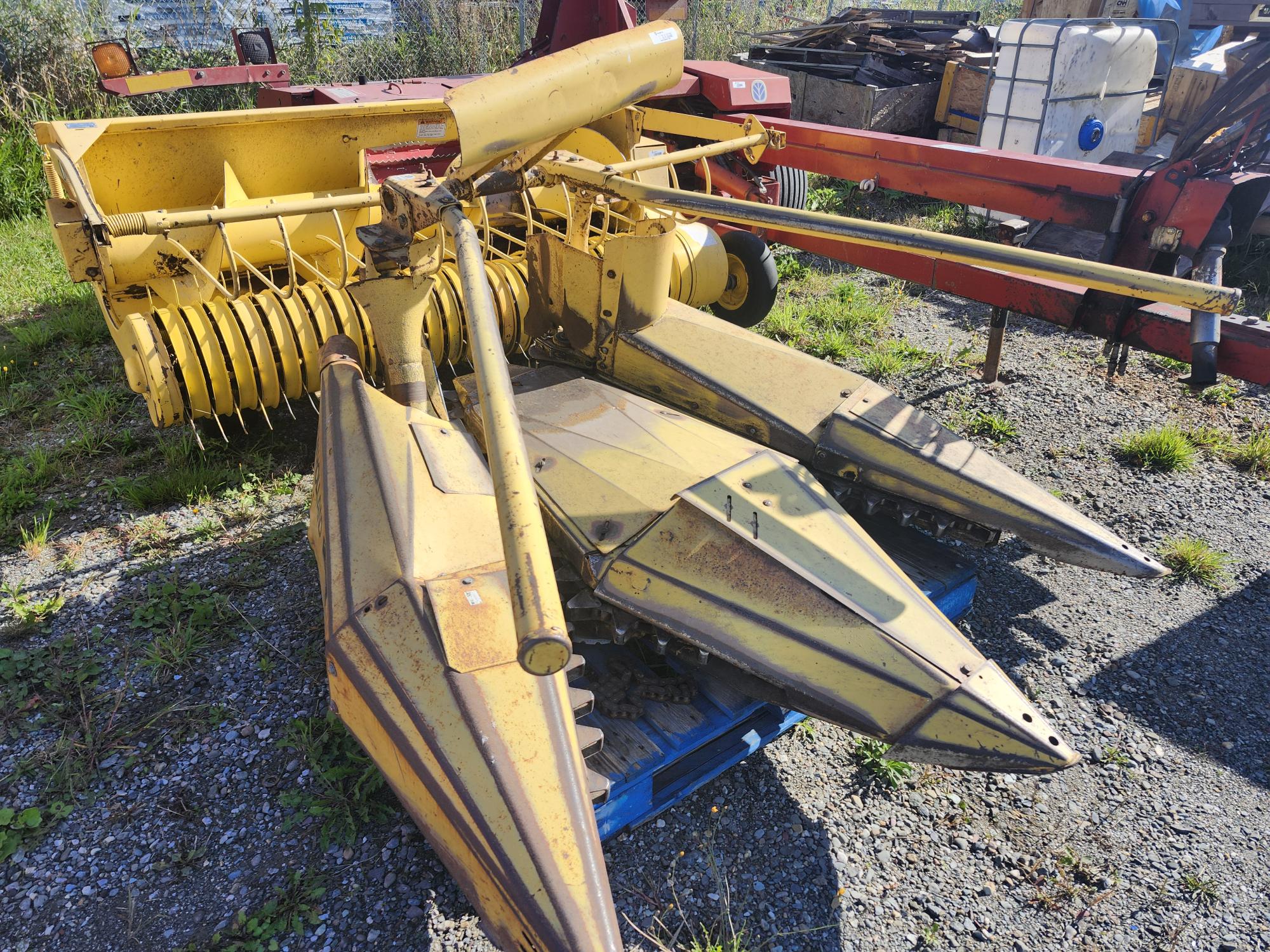 Une machine agricole jaune et rouillée, dotée de plusieurs pièces métalliques pointues, est posée sur le sol à l'extérieur. Elle est entourée d'herbe et de machines, et la zone est éclairée par la lumière du soleil.