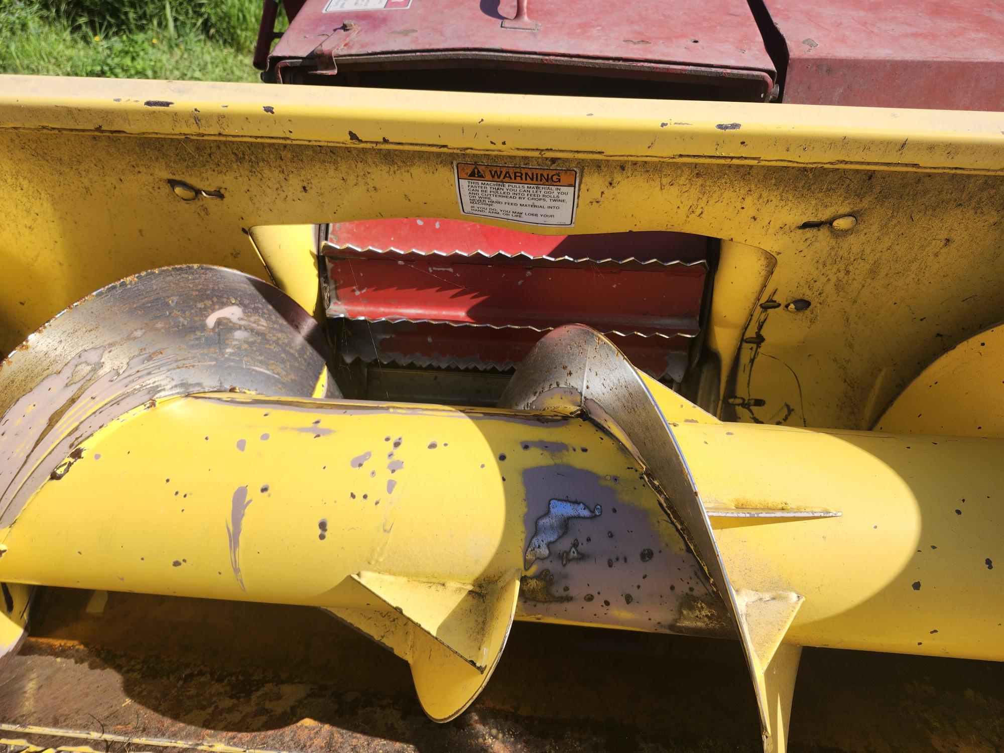 Gros plan d'un composant de machine agricole jaune avec des lames métalliques et un fond rouge. Une étiquette d'avertissement est apposée près du haut. La surface métallique est usée et légèrement rouillée.