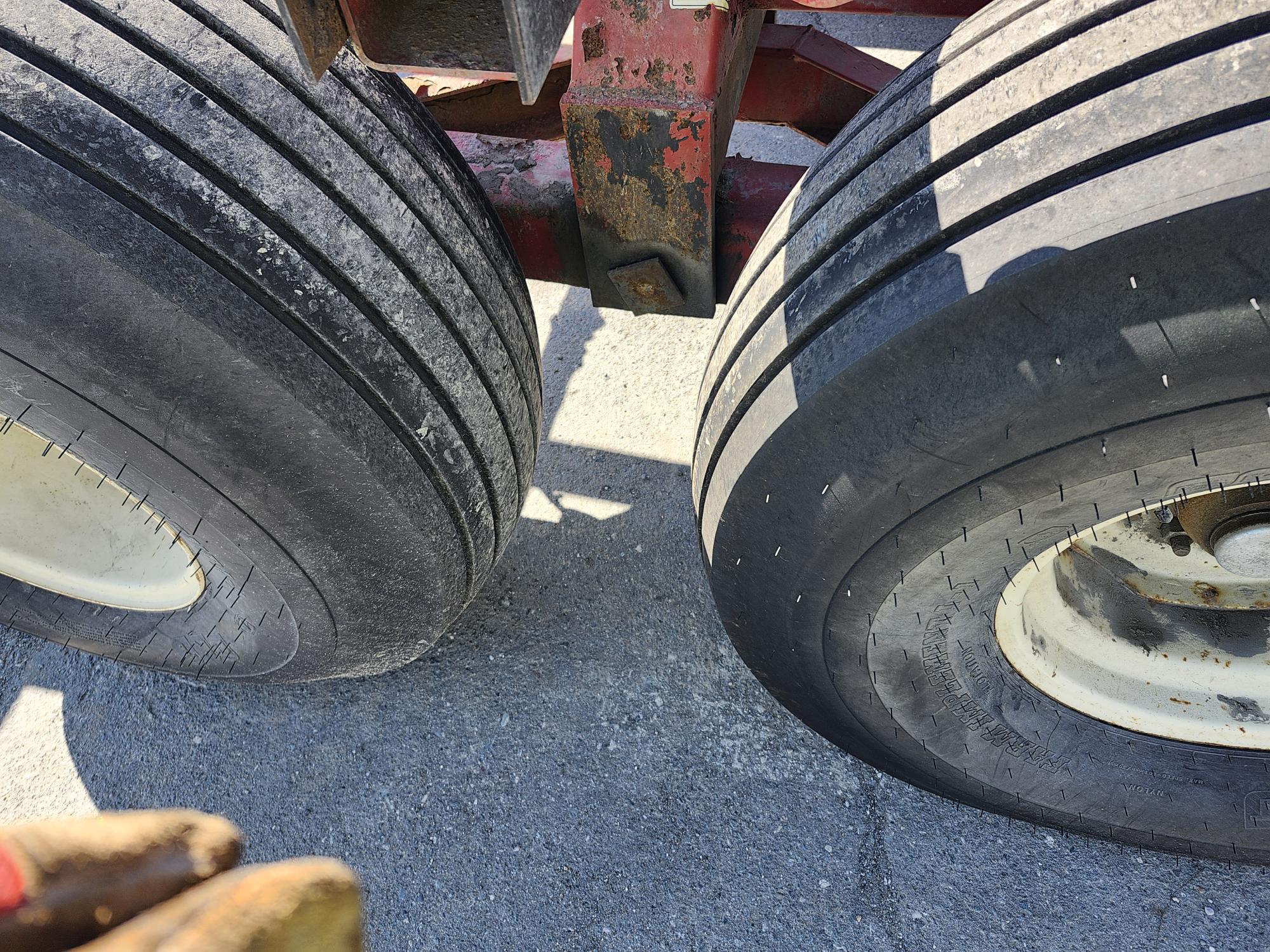 Gros plan de deux gros pneus sur l'essieu arrière d'un véhicule, posés sur une surface asphaltée. L'image montre les sculptures de la bande de roulement et les détails du caoutchouc. À proximité, une partie d'une main gantée est visible dans le coin inférieur gauche.