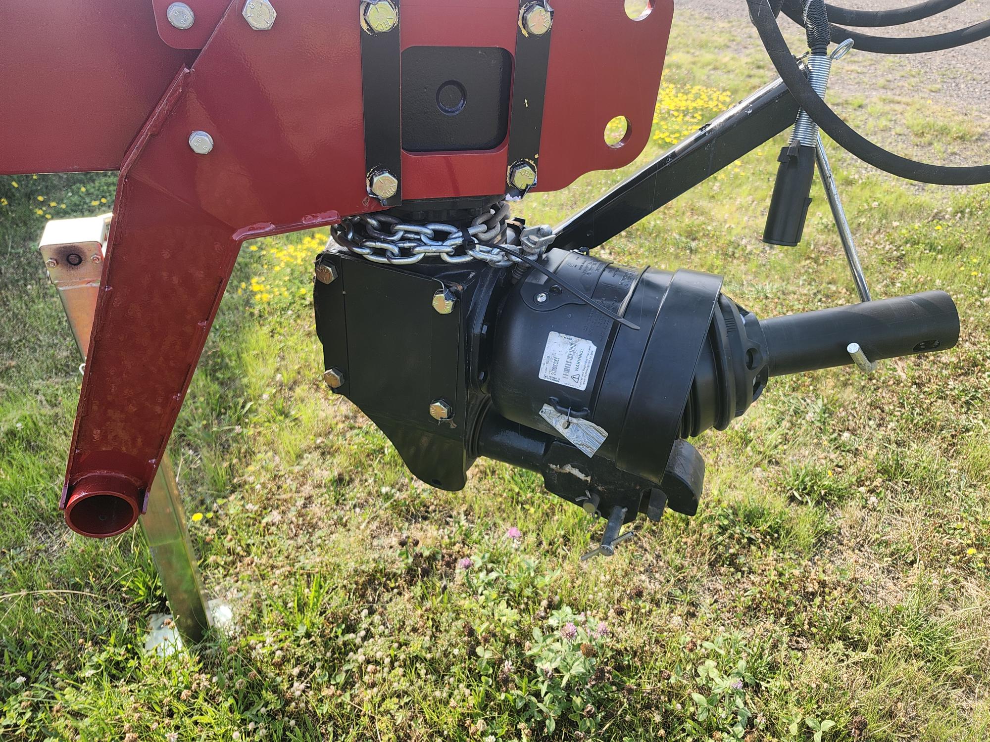 Gros plan d'un composant mécanique noir fixé à une machine agricole rouge, située sur un terrain herbeux. Le composant est maintenu en place par une chaîne et présente un câblage visible.