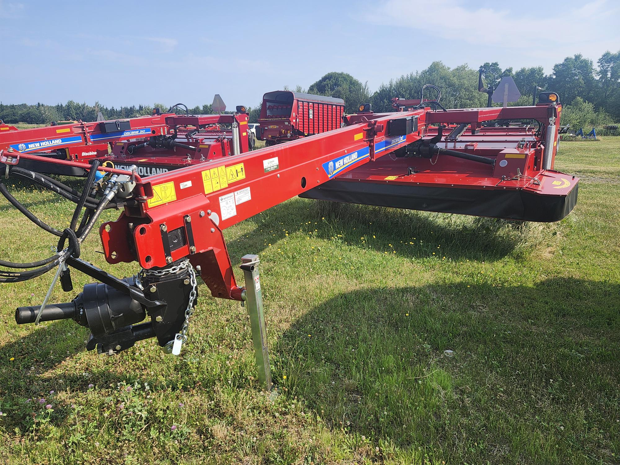 Une machine agricole rouge, probablement une faucheuse ou une presse à balles, est garée sur un terrain herbeux. Elle comporte diverses pièces mécaniques et des étiquettes de marque, avec des arbres et un ciel bleu visibles en arrière-plan.