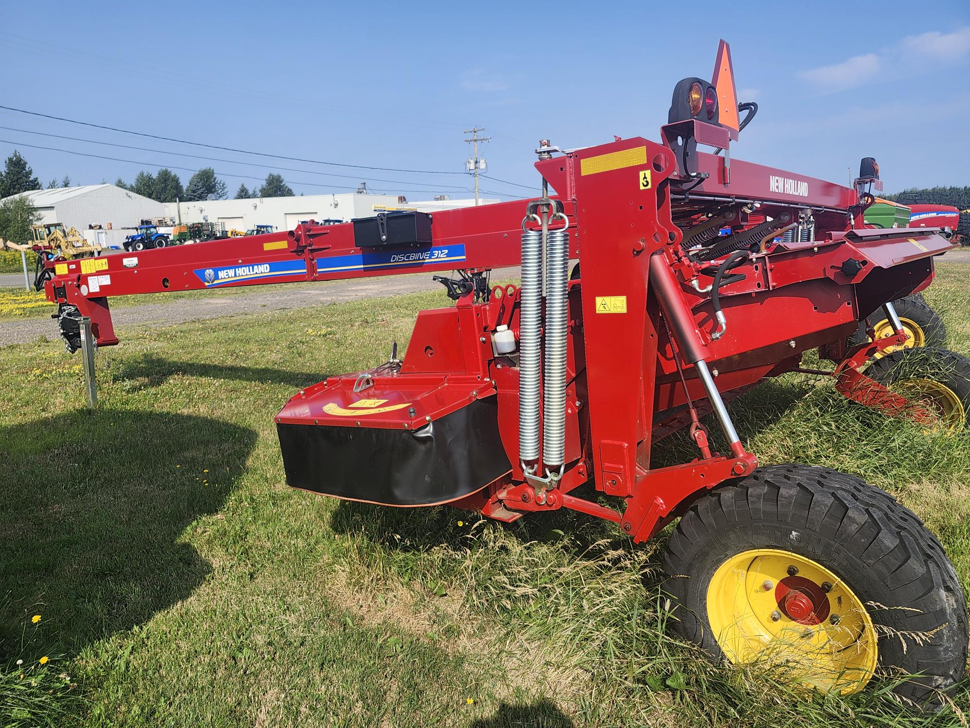 Une grande machine agricole rouge avec le logo « New Holland » est garée sur un terrain herbeux. Elle est dotée de roues jaunes et d'un mécanisme complexe. Des bâtiments et un ciel bleu clair sont visibles en arrière-plan.