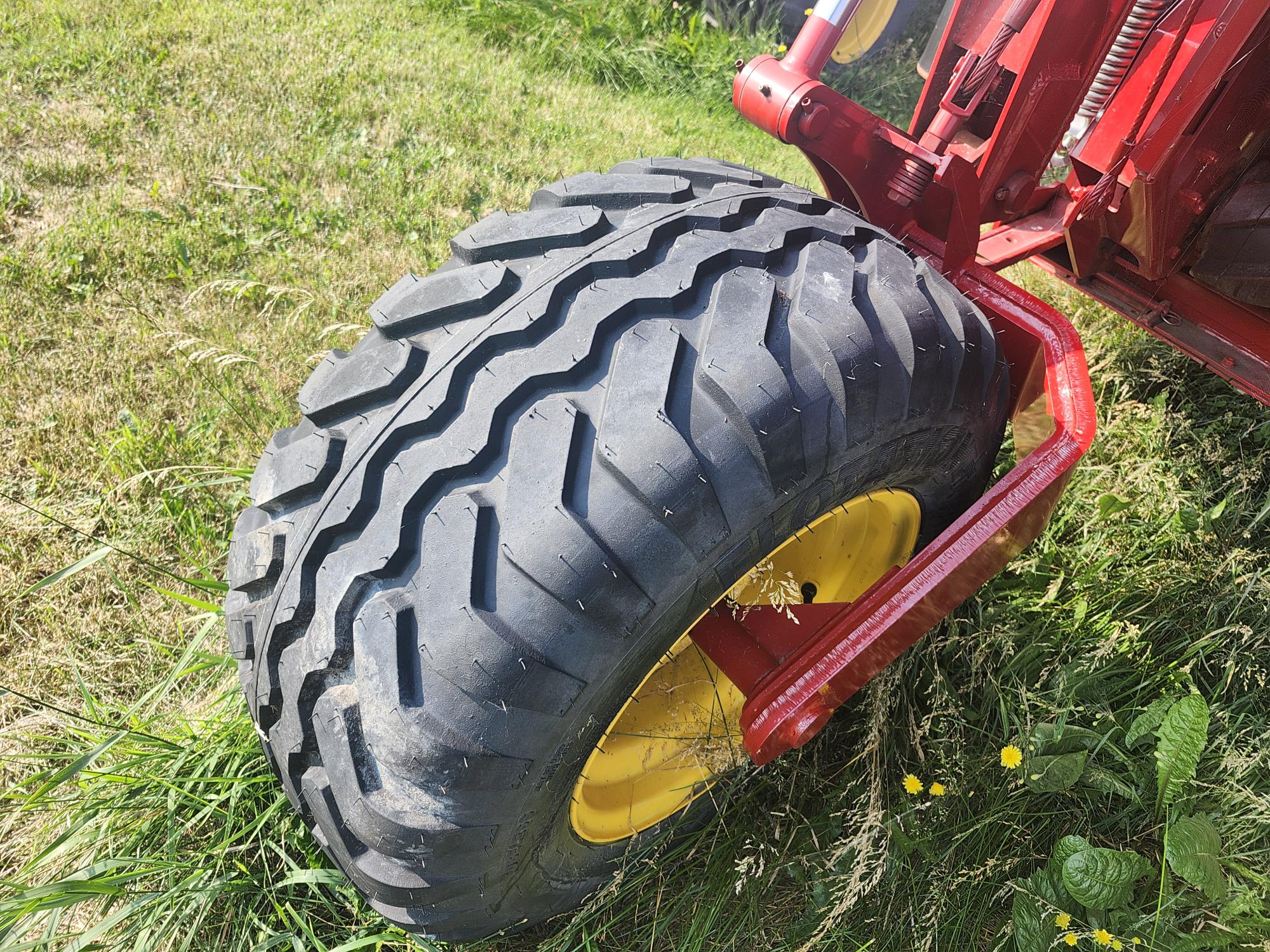 Un gros pneu de tracteur avec une jante jaune est partiellement encastré dans l'herbe. La structure métallique rouge du tracteur est visible au-dessus du pneu. La zone est entourée d'herbe verte luxuriante et de quelques petites fleurs jaunes.