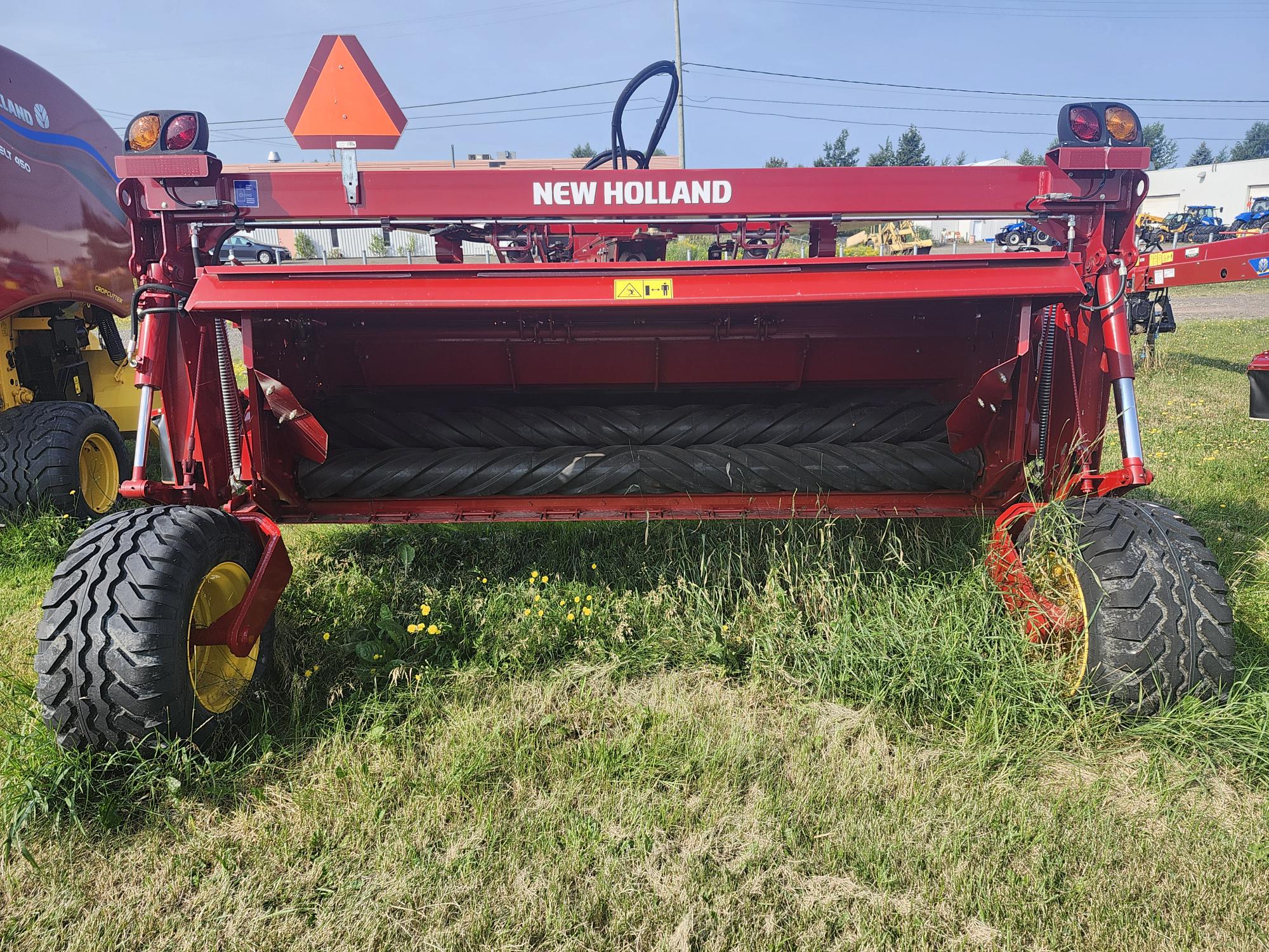 Une machine agricole New Holland rouge est garée sur une zone herbeuse avec de gros pneus et un mécanisme à courroie. Elle se détache sur un arrière-plan composé d'autres équipements agricoles et d'un ciel dégagé.