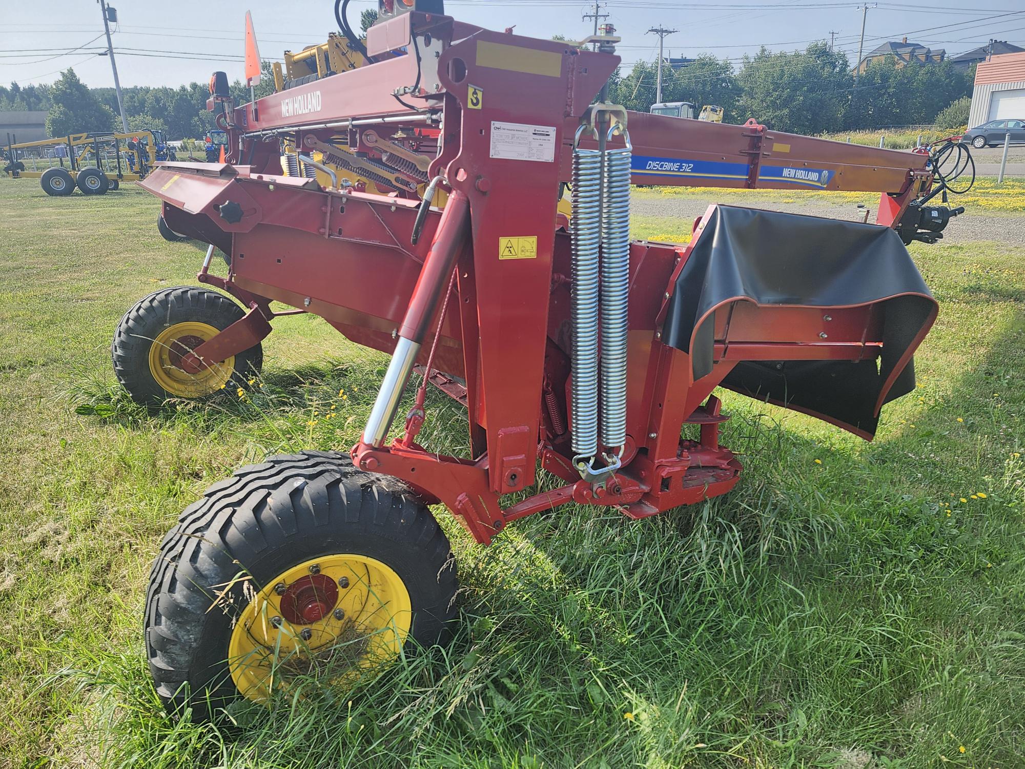 Machine agricole rouge avec roues jaunes sur un terrain herbeux. L'équipement comprend de gros ressorts et des bras métalliques. L'arrière-plan présente des arbres, des bâtiments et un ciel clair.