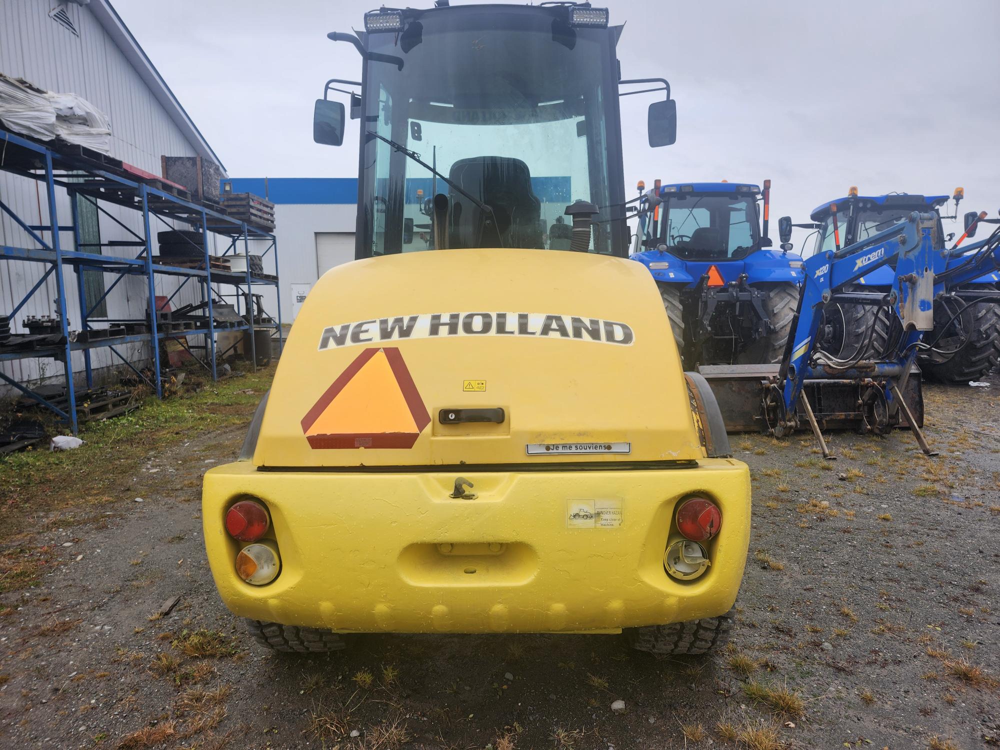 Vue arrière d'un véhicule de chantier New Holland jaune stationné à l'extérieur sur une surface en gravier. Il est doté d'un panneau triangulaire rouge et orange et de deux feux rouges. Des tracteurs bleus sont alignés à l'arrière-plan près d'un bâtiment blanc et bleu.