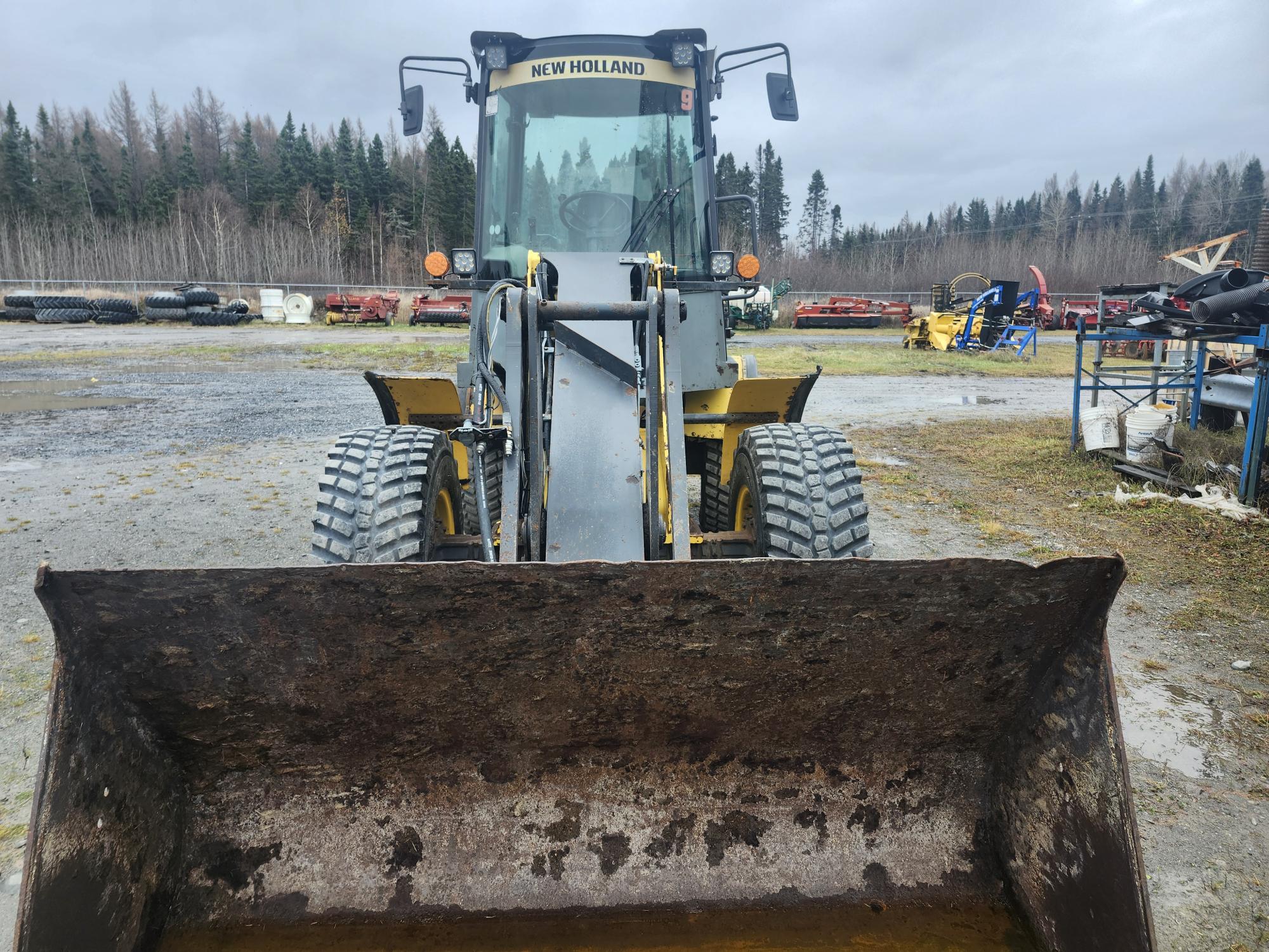 Vue de face d'une chargeuse New Holland jaune avec un godet rouillé, garée sur une surface en gravier. En arrière-plan, il y a des arbres et diverses pièces de machines éparpillées dans une cour sous un ciel nuageux.