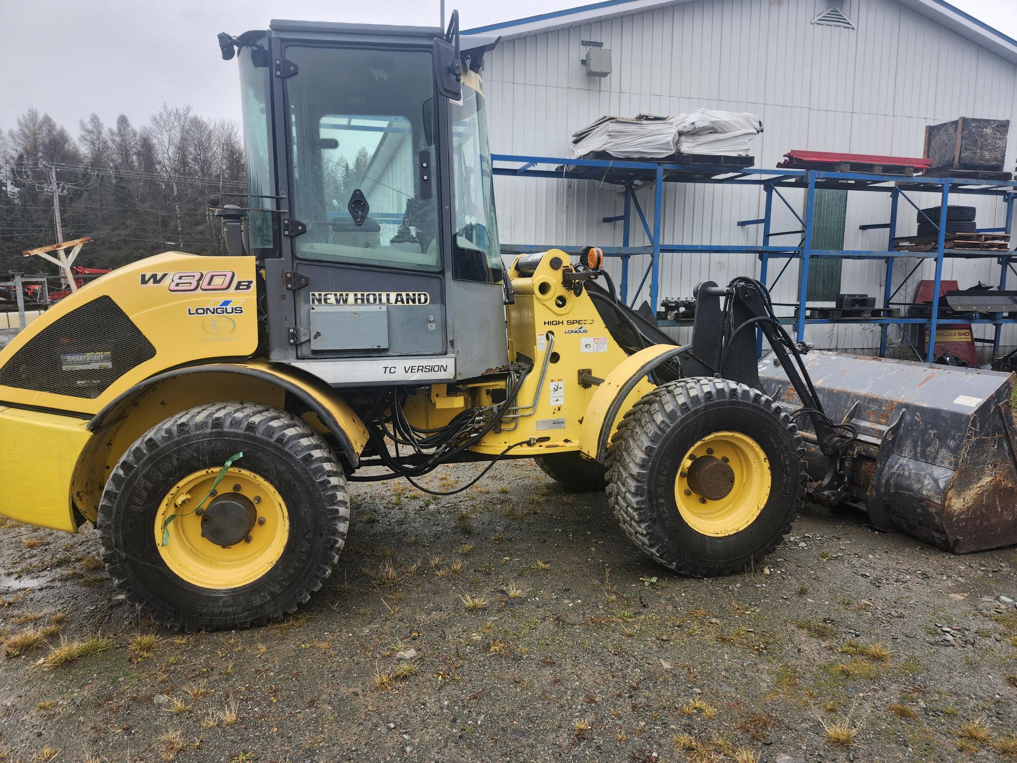 Une chargeuse sur pneus jaune New Holland W80B TC est garée sur du gravier devant un bâtiment industriel avec des racks métalliques. Ciel couvert et arbres en arrière-plan.