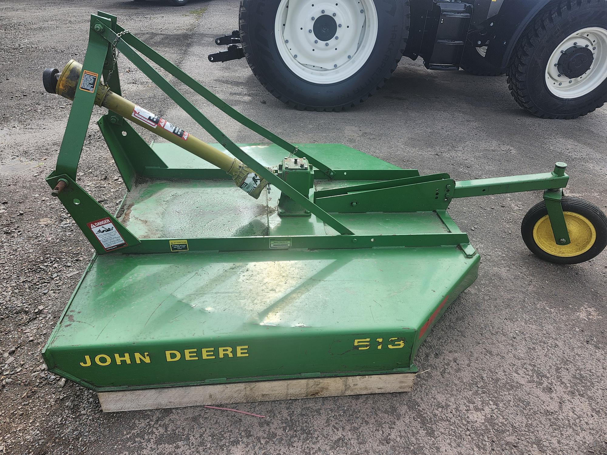 Une fraise rotative verte John Deere 513 est présentée sur une surface en gravier, avec un véhicule plus grand en arrière-plan. La fraise est dotée d'une roue jaune et est légèrement usée, ce qui indique son utilisation.