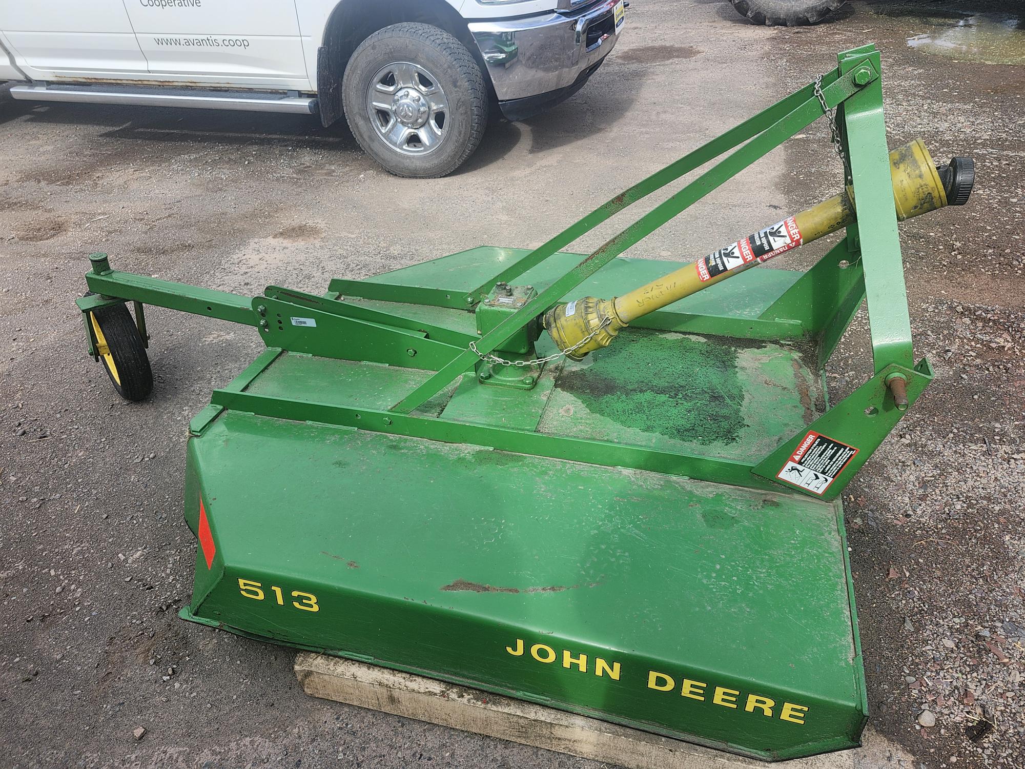 Un accessoire de coupe rotatif vert John Deere étiqueté « 513 » est stationné sur une surface en terre. Un véhicule blanc et d'autres équipements sont partiellement visibles à l'arrière-plan.